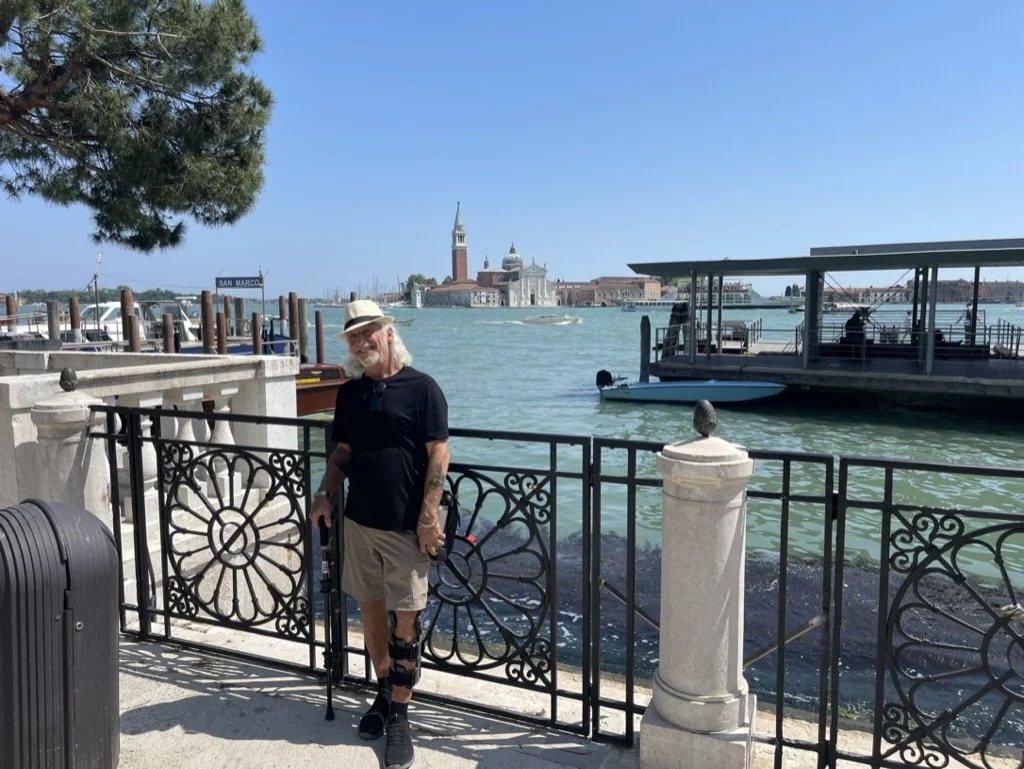 An elderly man with a hat, sunglasses, black shirt, shorts, and a cane standing by a decorative metal gate near a waterfront, with boats on the water and historic buildings in the background.