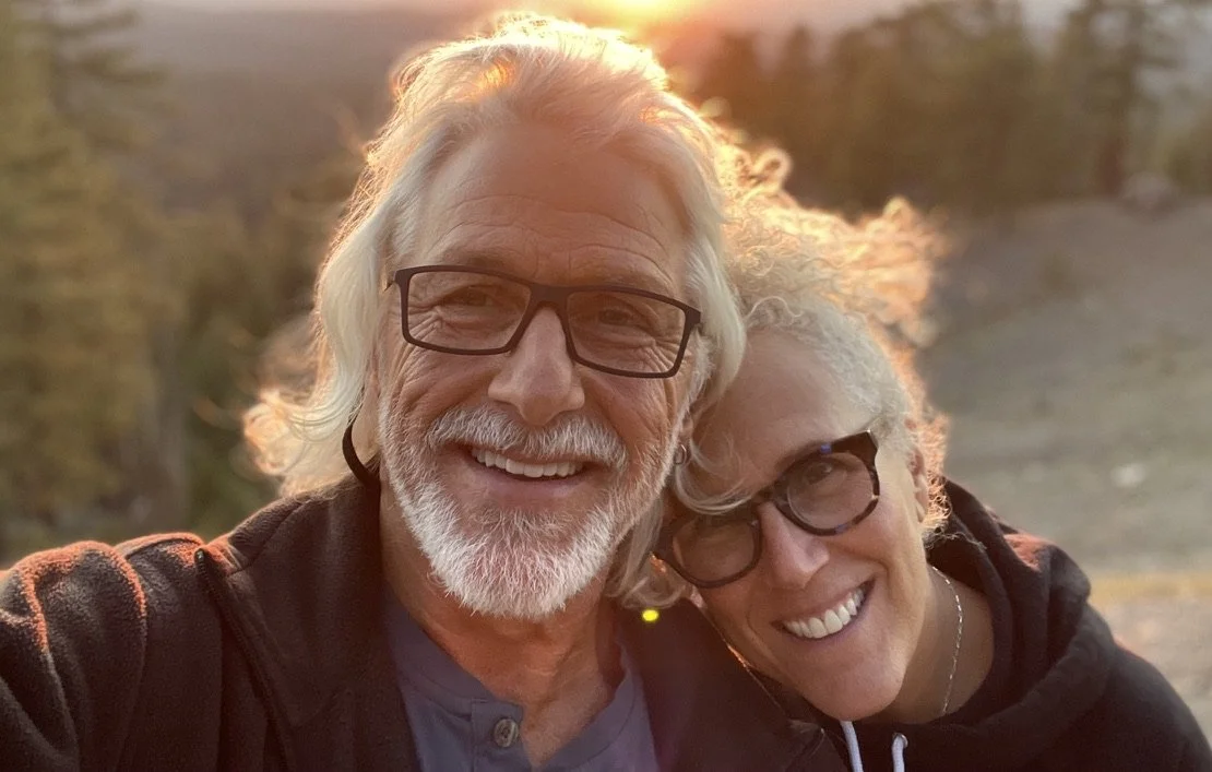 A happy older man and woman smiling, wearing glasses outdoors during sunset with trees in the background.