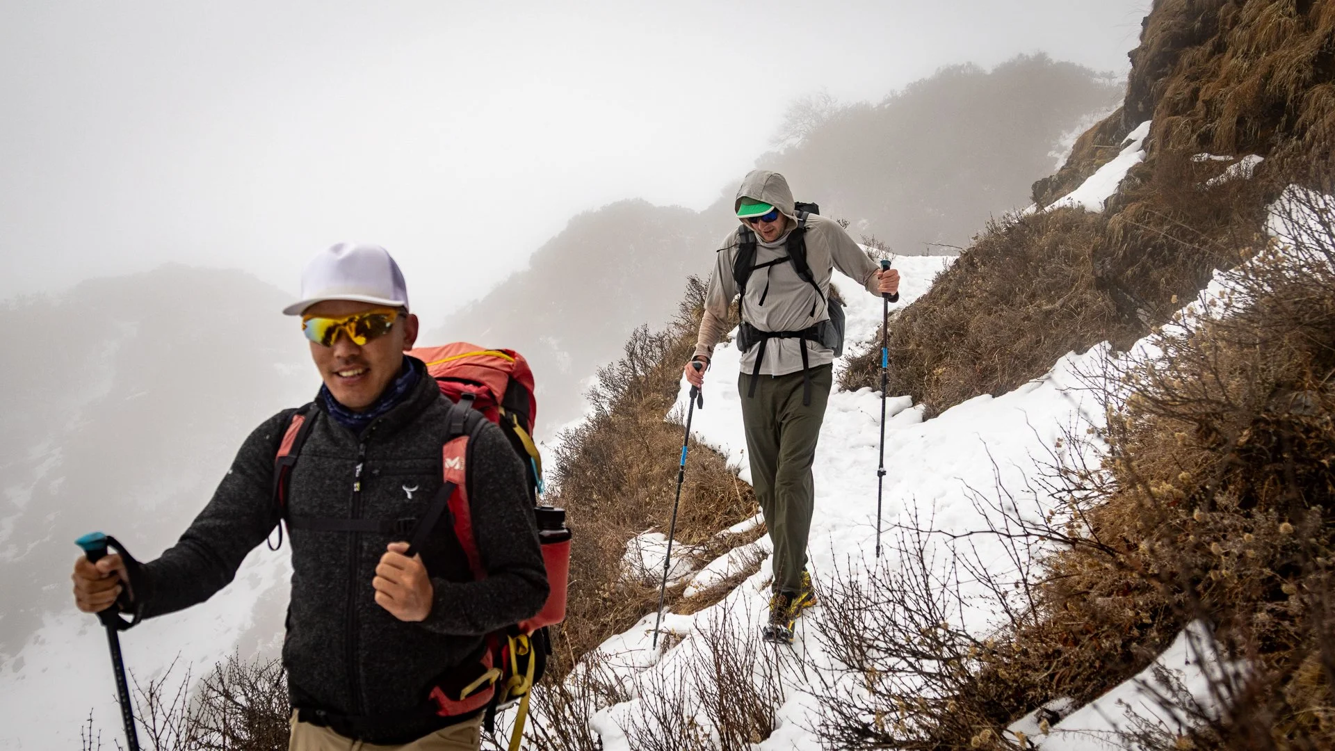 Two hikers trekking through a snowy mountain trail with foggy conditions and sparse bushes.