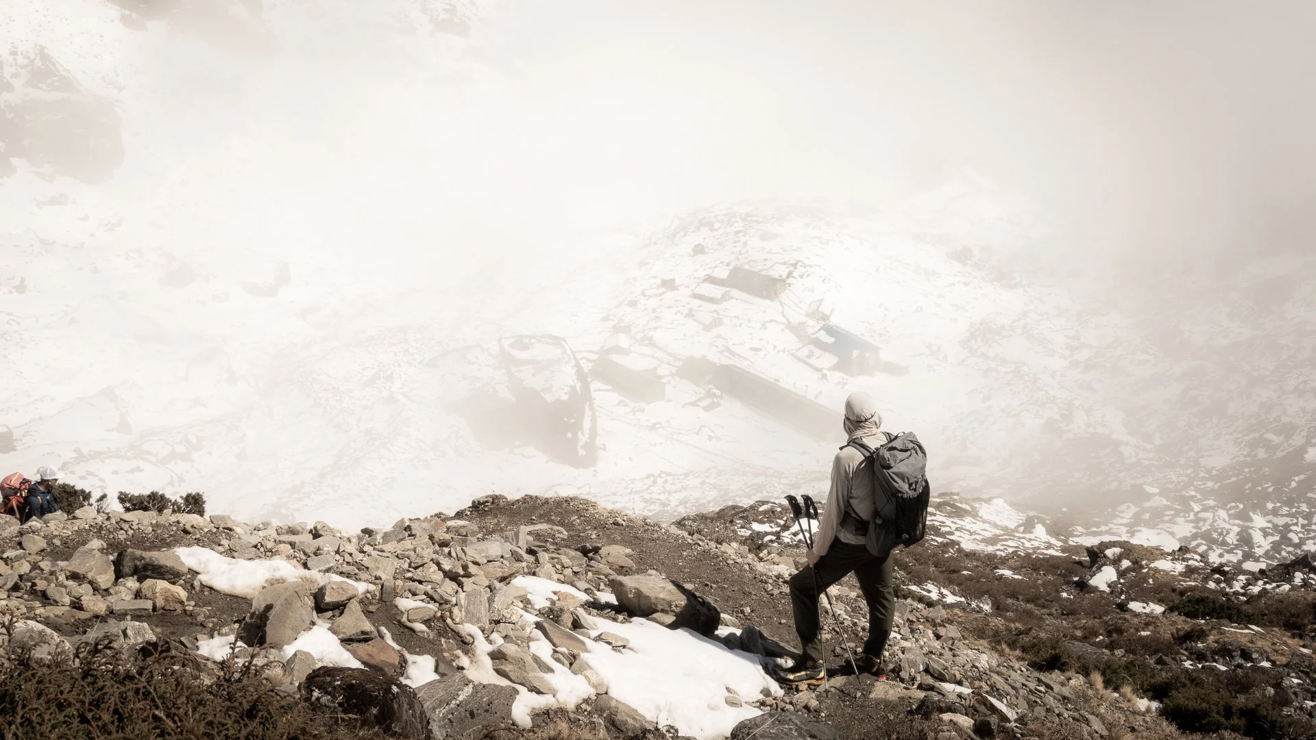 Hiker in gray outdoor gear and backpack standing on rocky snowy mountain slope, overlooking snow-covered mountain landscape with fog and debris below.