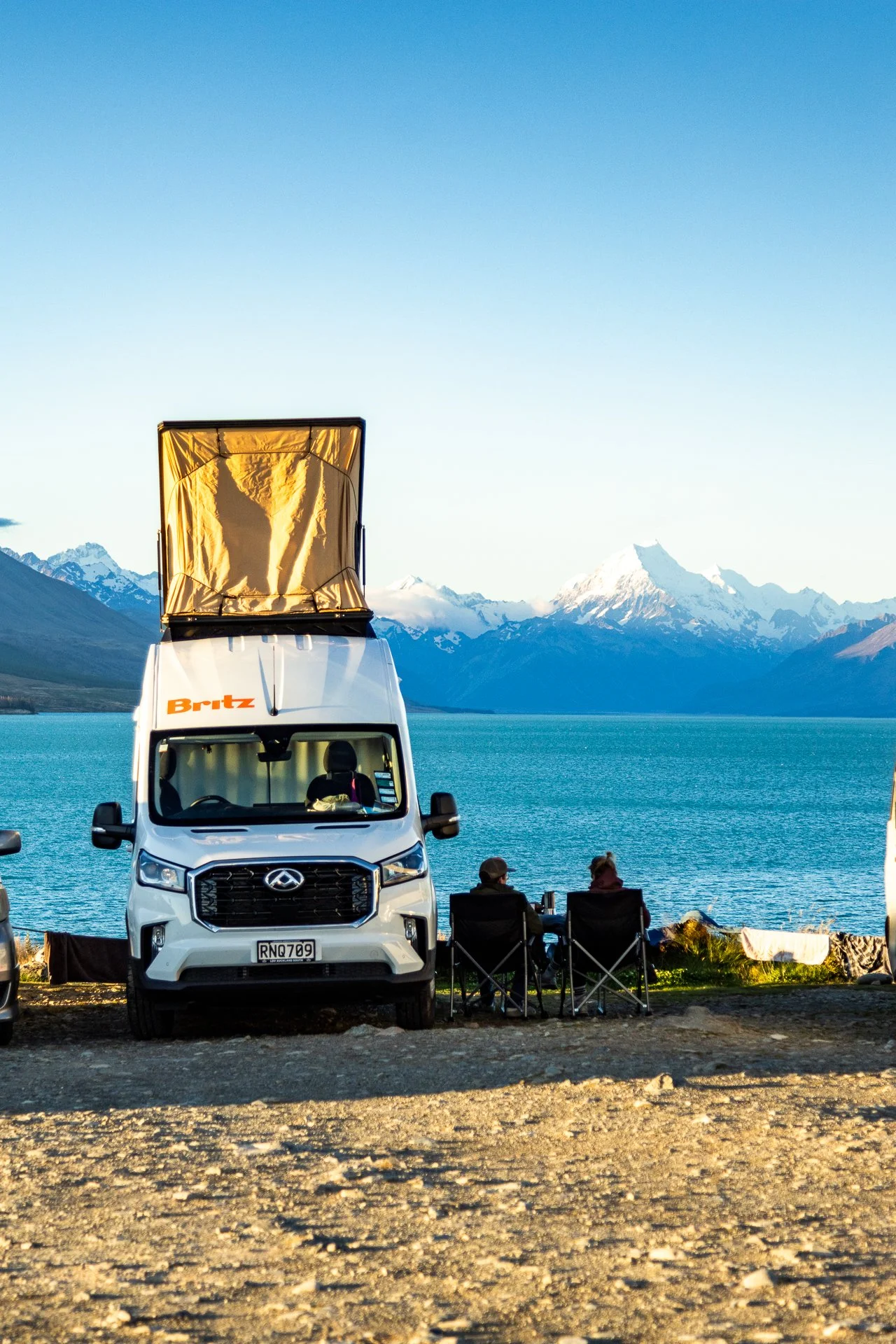A white camper van parked on a gravel area next to a lake, with two people sitting in camping chairs, mountains and snow-capped peaks in the background under a clear blue sky.