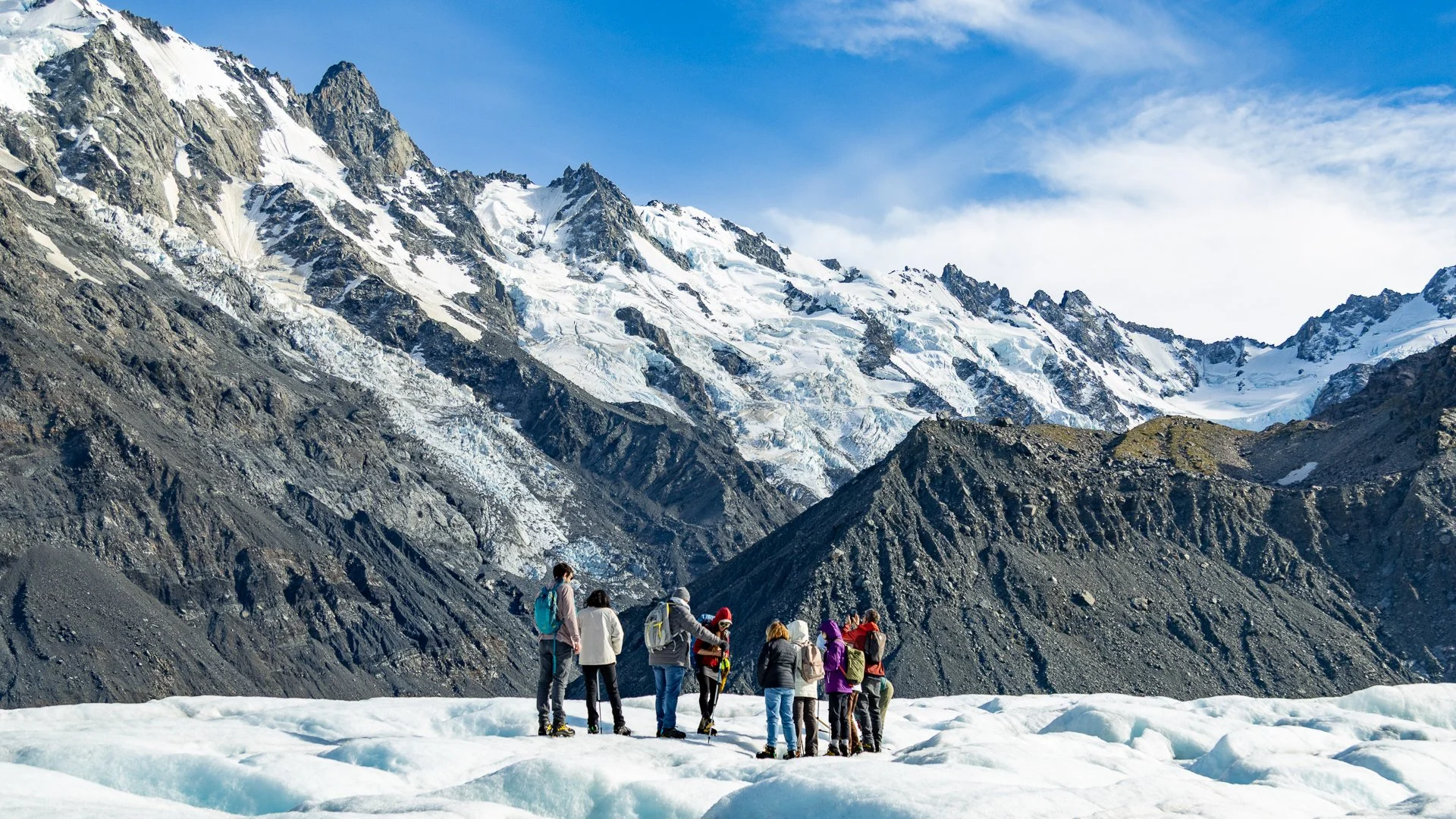 Group of hikers in winter gear on snow-covered ground in a mountainous area with rugged, snow-capped peaks and a partly cloudy sky in the background.