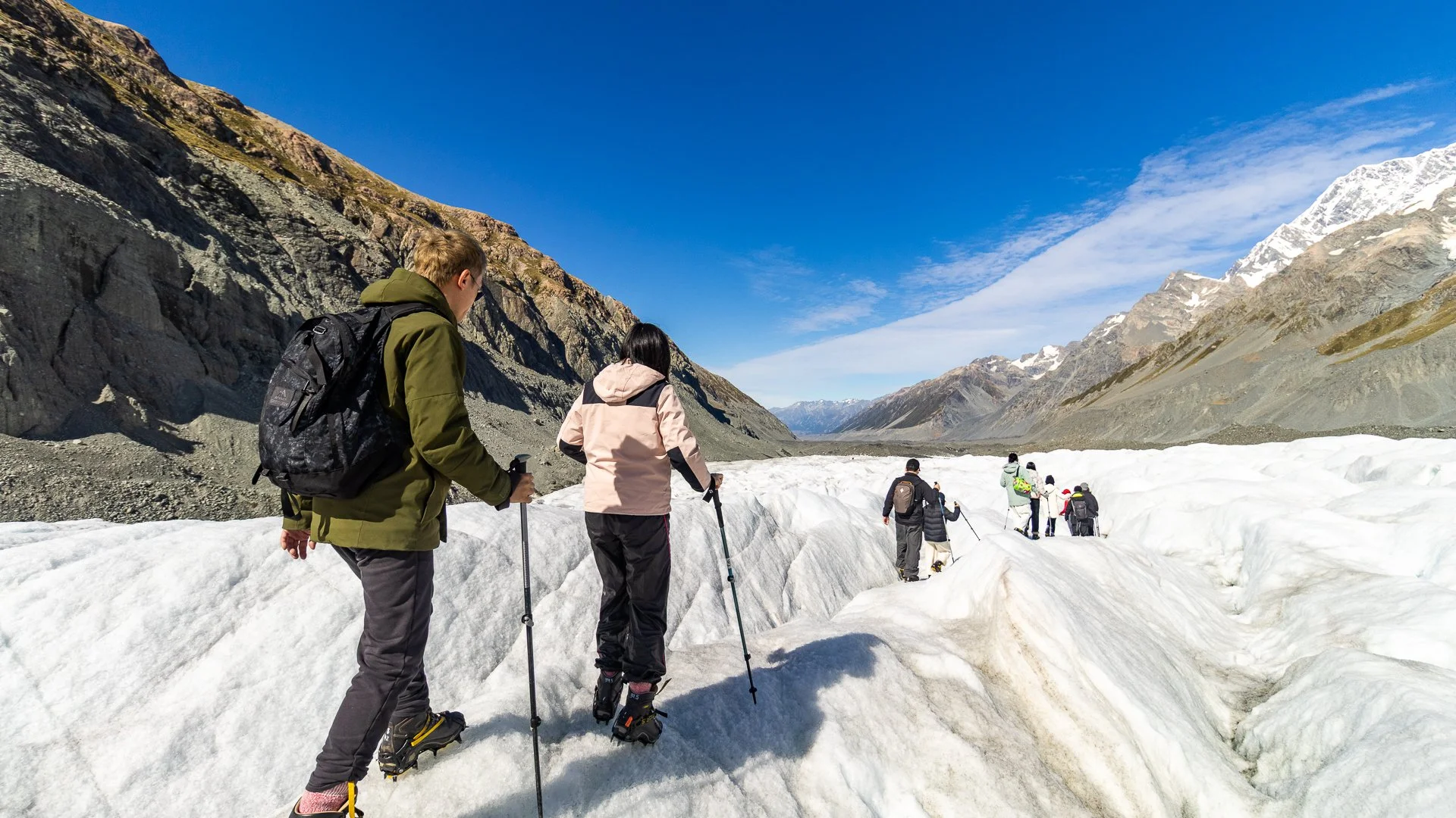 Group of people hiking on snowy terrain in a mountainous area with clear blue sky, snow-capped mountains, and rocky slopes in the background.