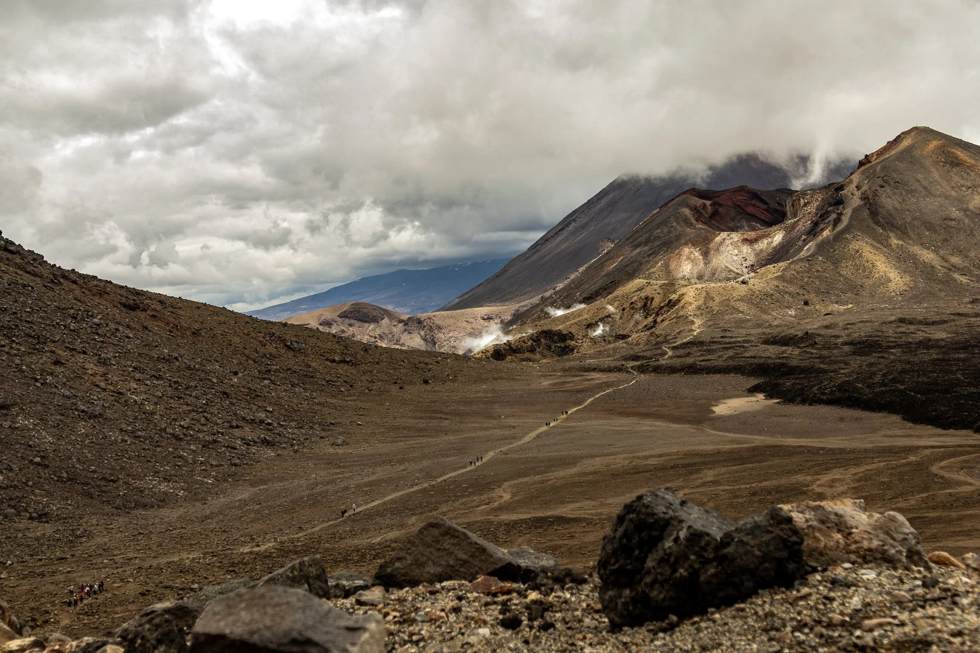 A volcanic landscape with barren, rocky terrain under a cloudy sky. A trail winds through the landscape with small groups of hikers visible in the distance.