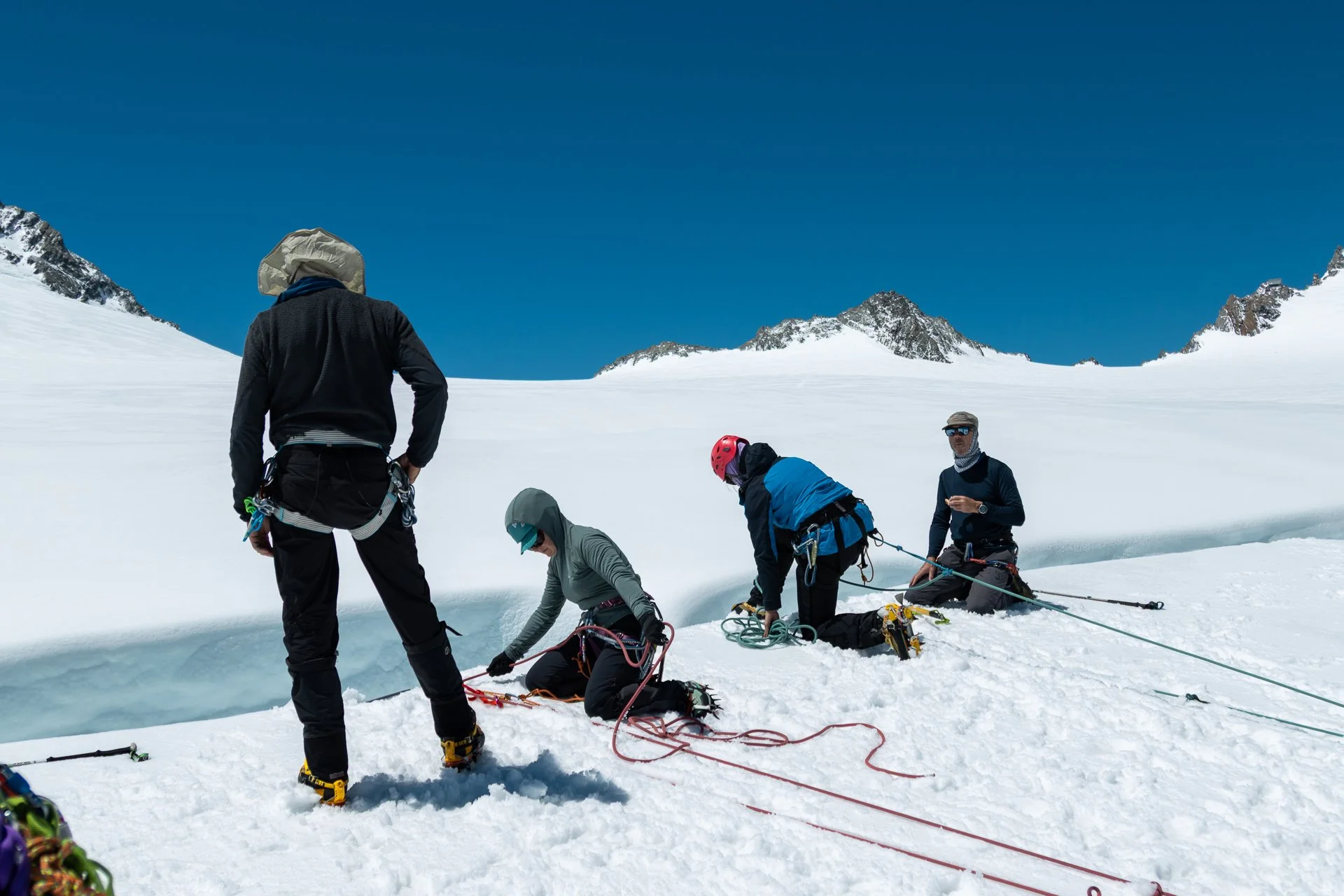 Group of climbers in snow-capped mountain landscape, some kneeling and some standing, preparing gear, under a clear blue sky.