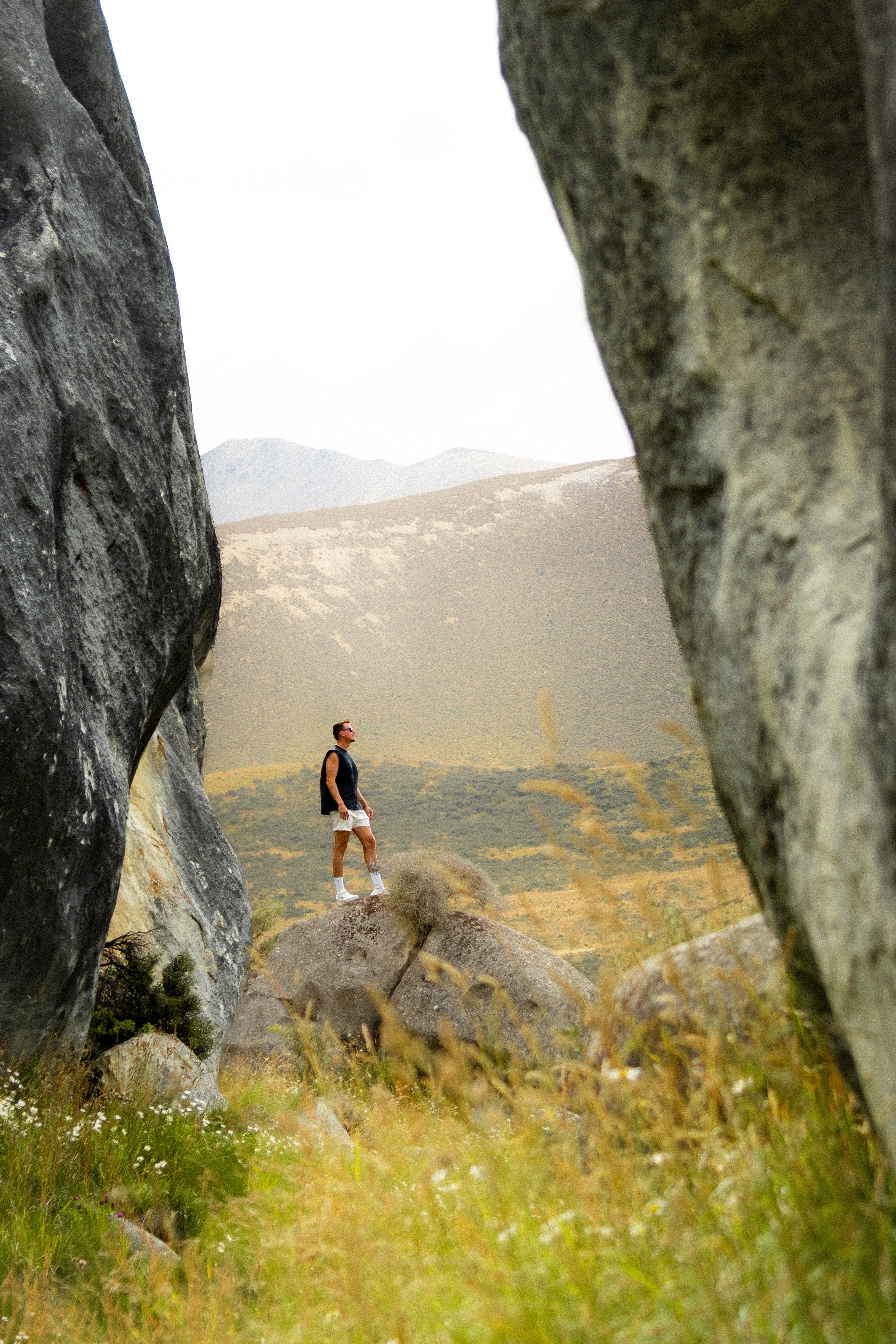 A man standing on a rock in a mountainous landscape, framed between two large rocks in the foreground, with hills and mountains in the background.