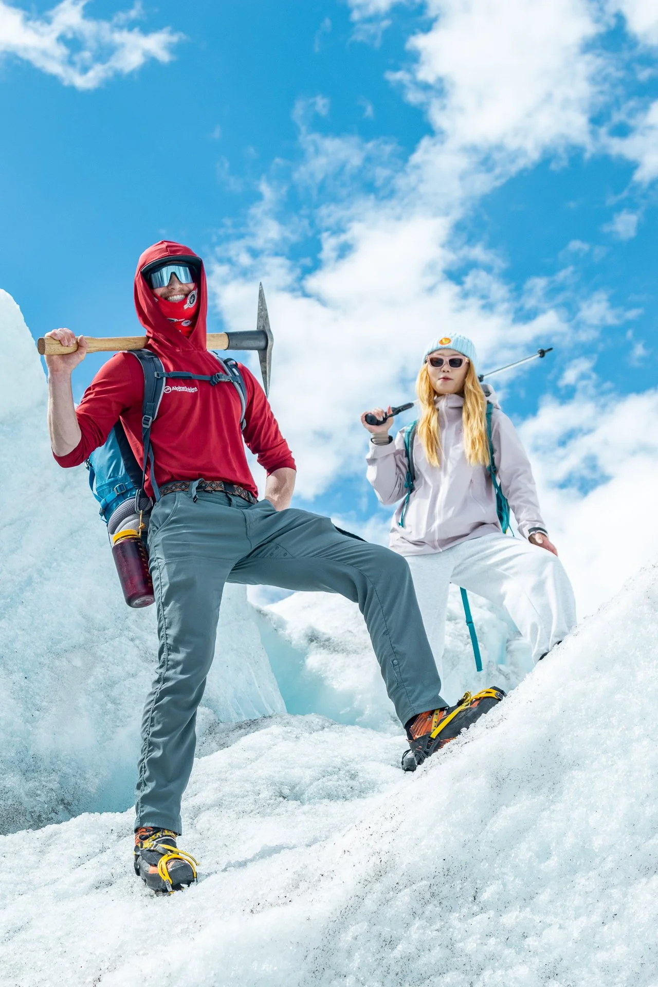 Two people dressed in snow gear, standing on snowy mountain terrain with blue sky and clouds overhead. One holds an ice axe, the other holds trekking poles.