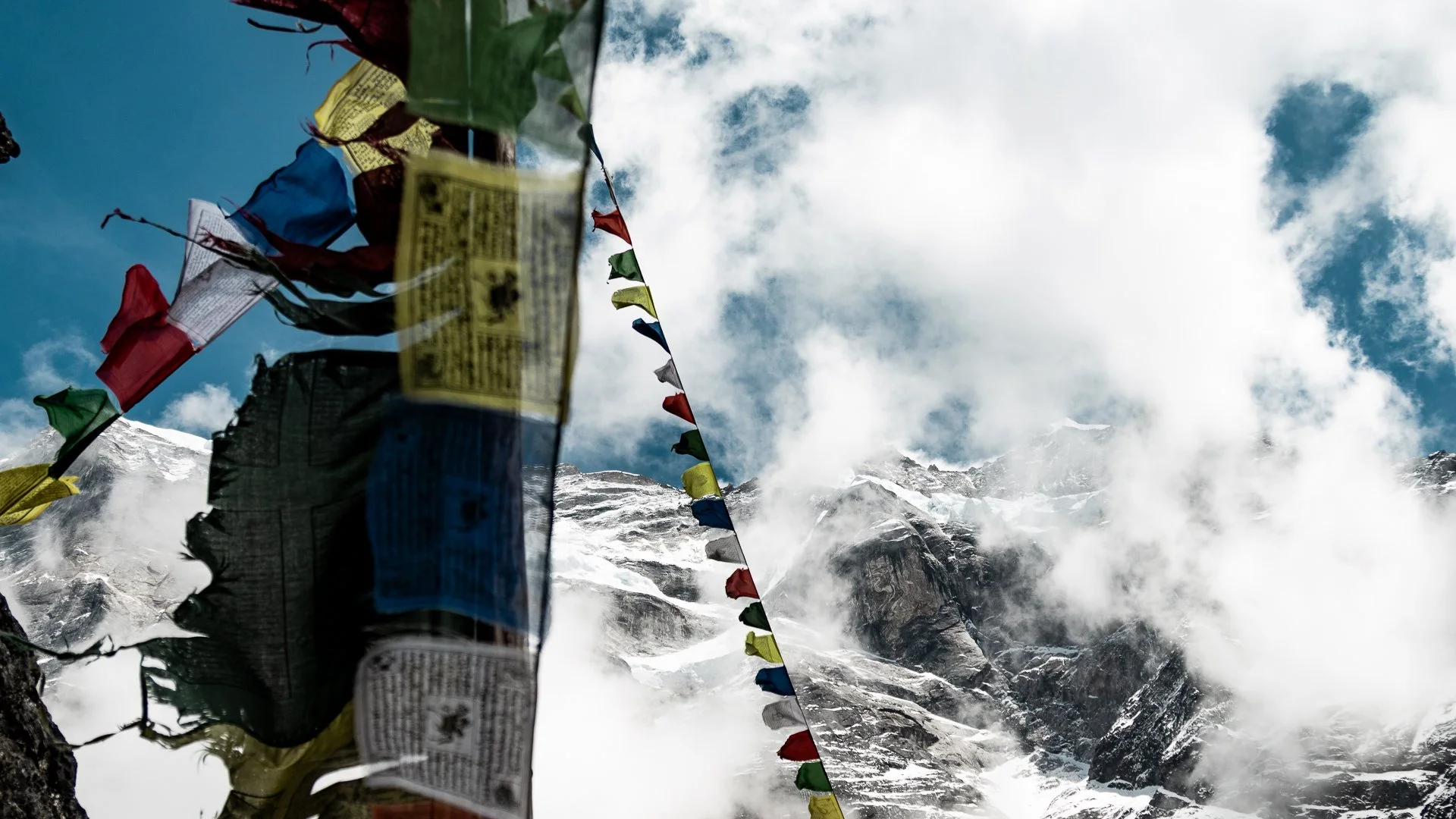 Prayer flags fluttering against a backdrop of snow-capped mountains and a partially cloudy sky.