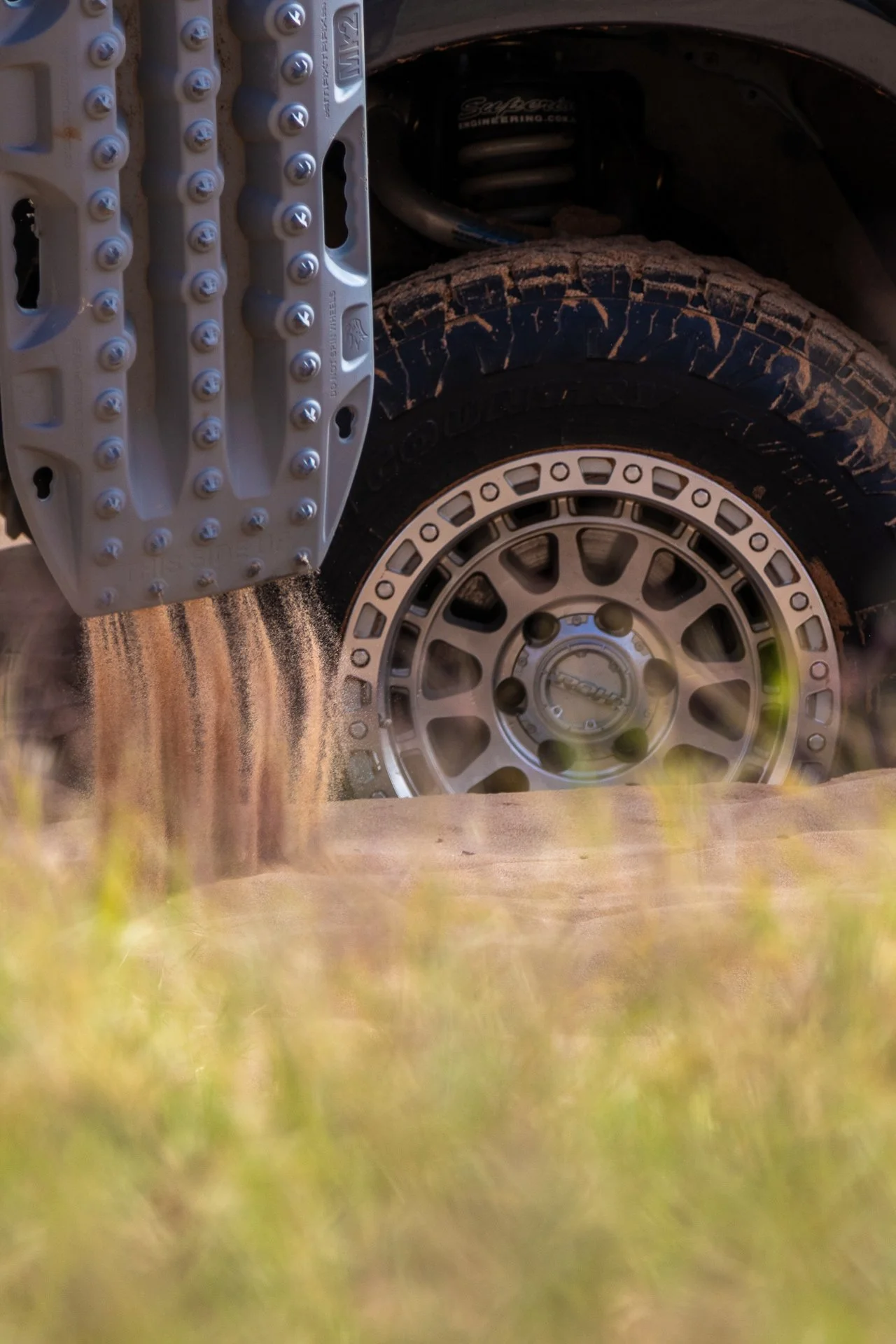 Close-up of a vehicle's tire with mud on the treads, and a metal wheel rim, partially obscured by grass in the foreground.