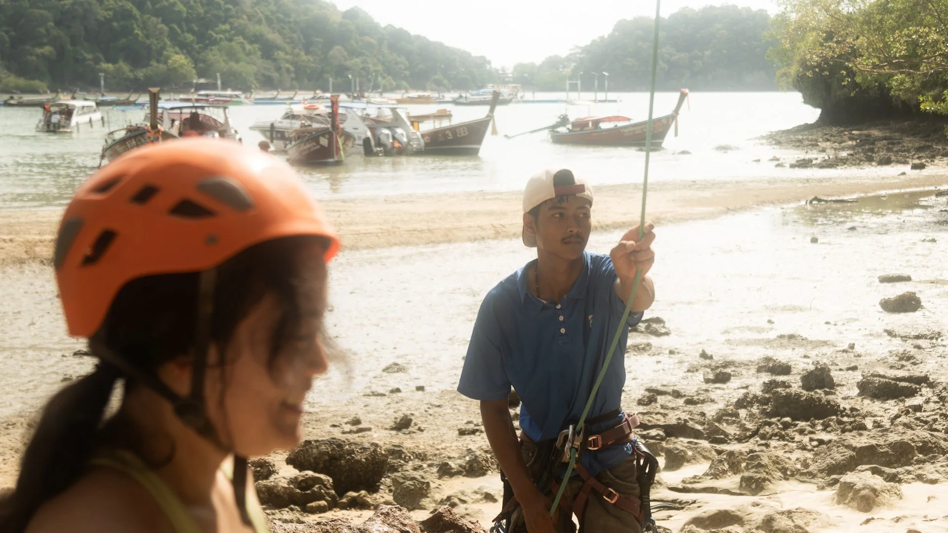 Two young individuals at a beach preparing for a water activity. One person in a yellow top and orange helmet facing away, and another person holding a rope, wearing a cap, blue shirt, and harness. Several boats are anchored in the water in the backg