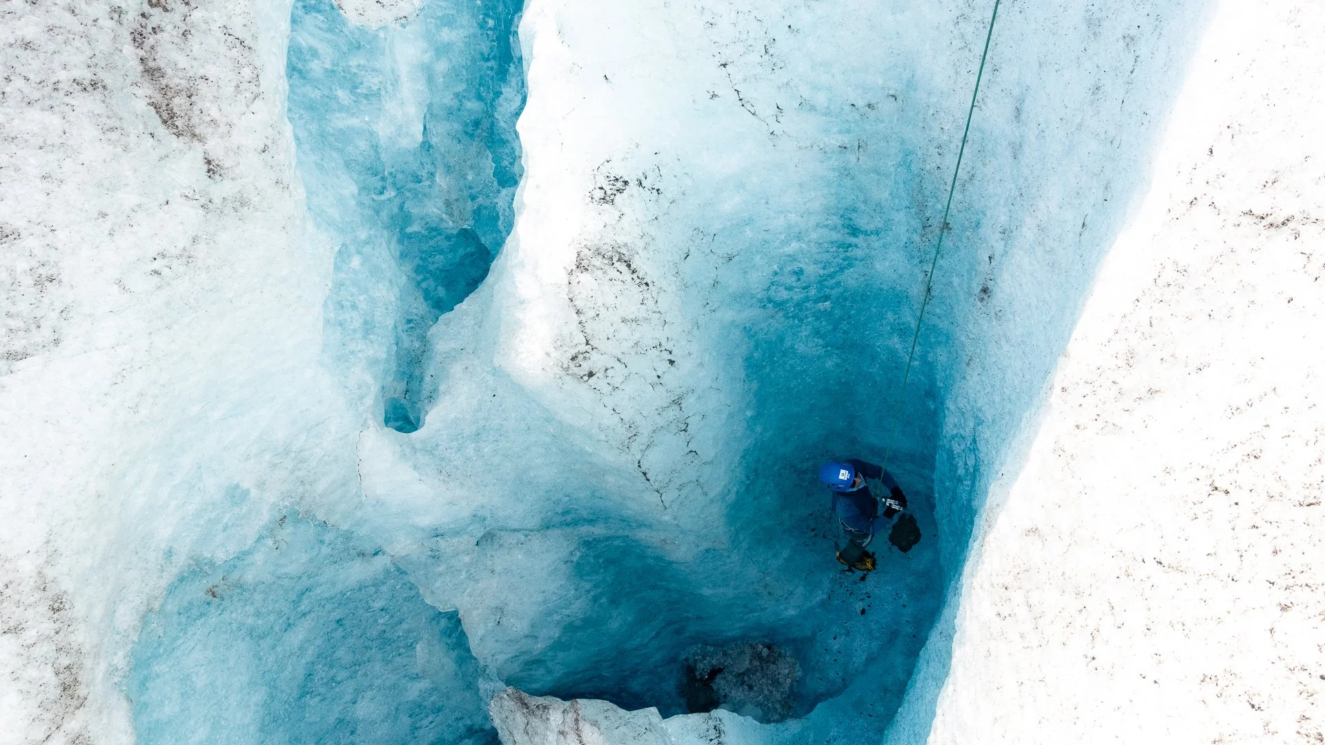 A person with a helmet inside a deep, blue ice crevasse in a glacier.