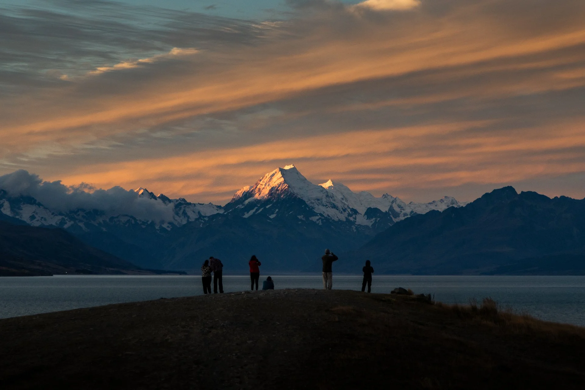 People standing on the shore observing snow-capped mountains at sunset, with orange and pink clouds in the sky.