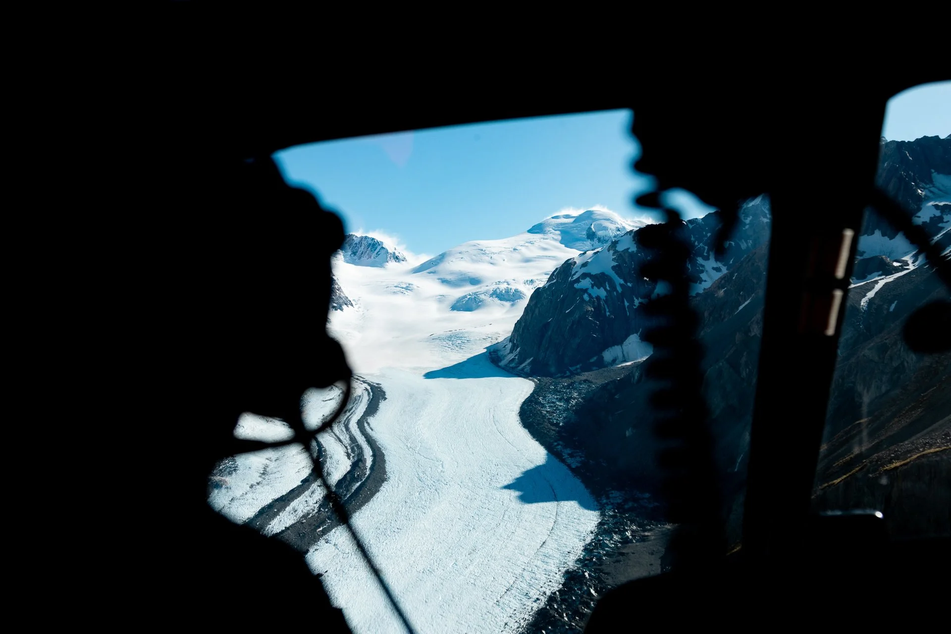 A view of snow-covered mountains and a glacier through the window of a helicopter or aircraft.