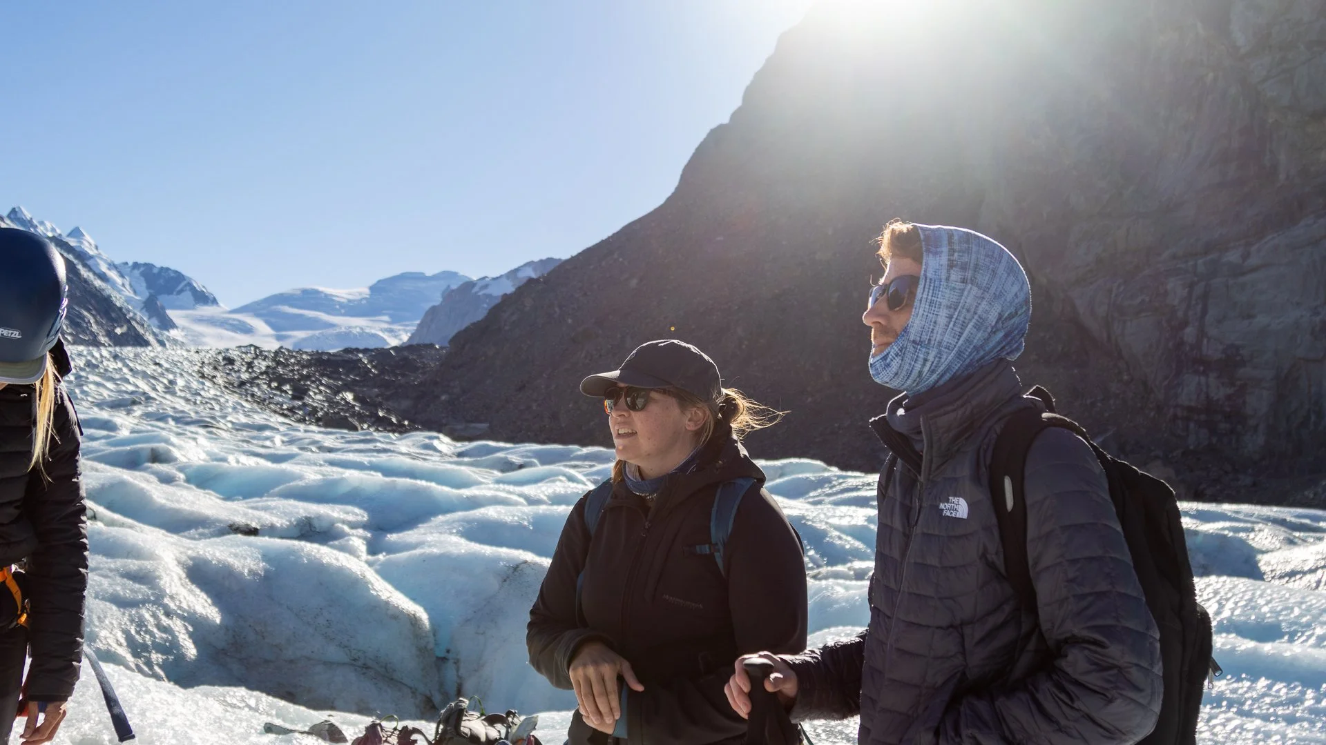 Group of hikers standing on snow and ice in a mountainous landscape with snow-capped peaks in the background and sunlight shining from above.
