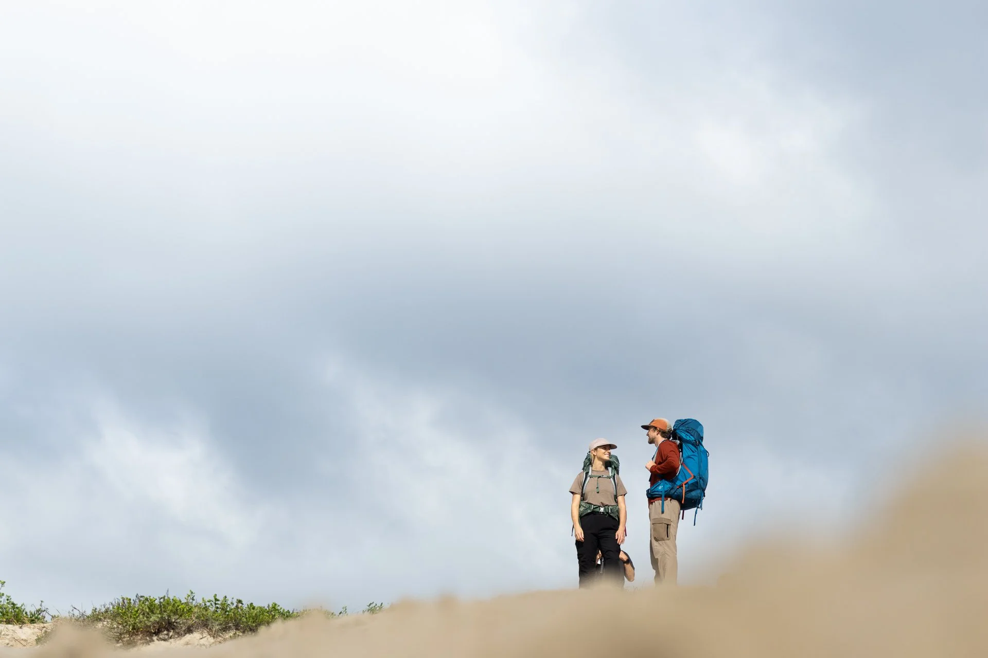 Two hikers with backpacks standing on a hill and talking, against a cloudy sky.
