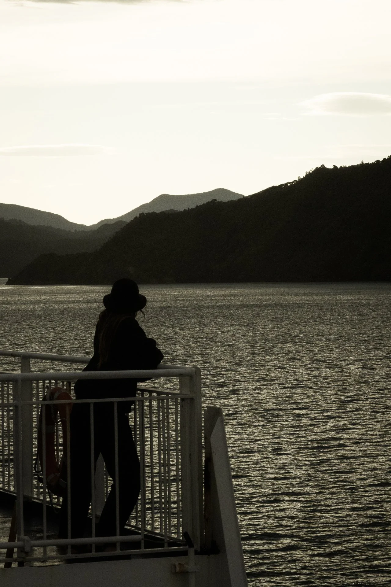 A person standing on a boat looking out over a body of water with mountains in the background during sunset or sunrise.