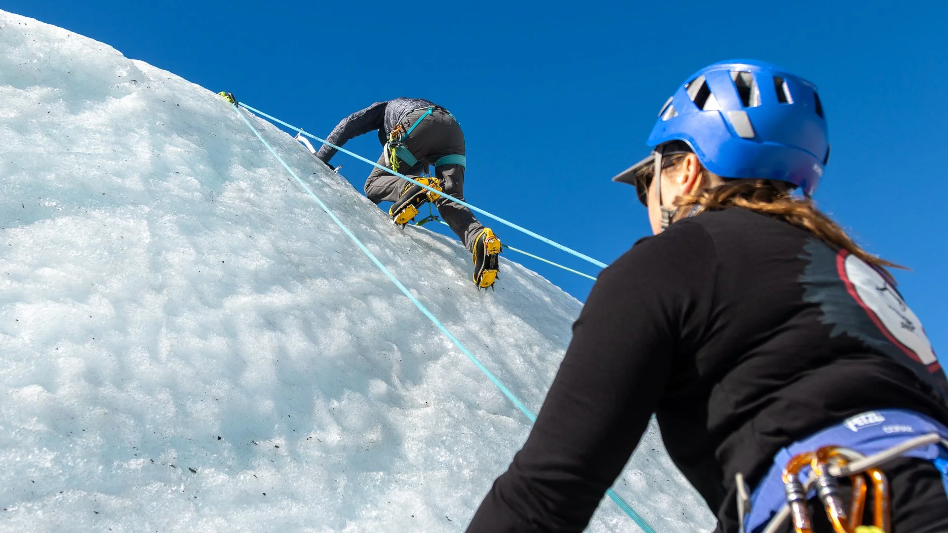 Two mountain climbers ascending an icy slope with safety ropes and helmets under a clear blue sky.