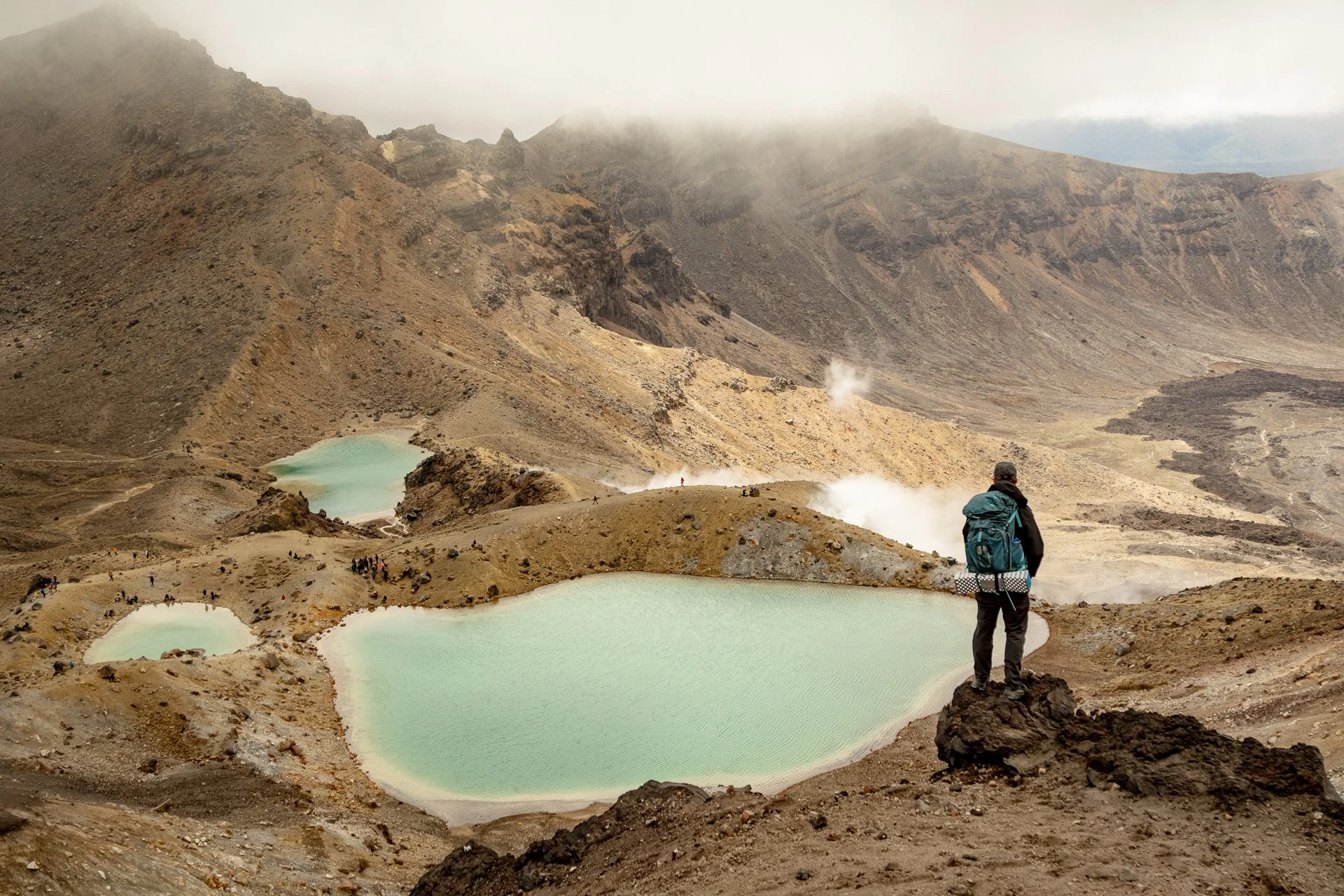 A hiker with a backpack standing on rocky terrain overlooking a volcanic crater with steaming vents and pale blue-green lakes, surrounded by mountains and fog.