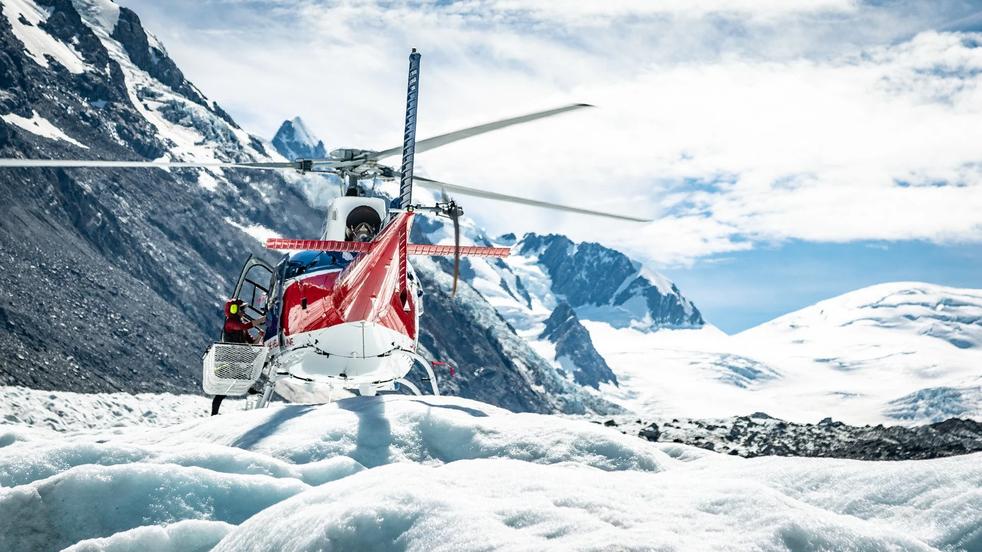A red and white helicopter on snow-covered ground in a mountainous, snowy landscape with glaciers and snow-covered peaks under a partly cloudy sky.