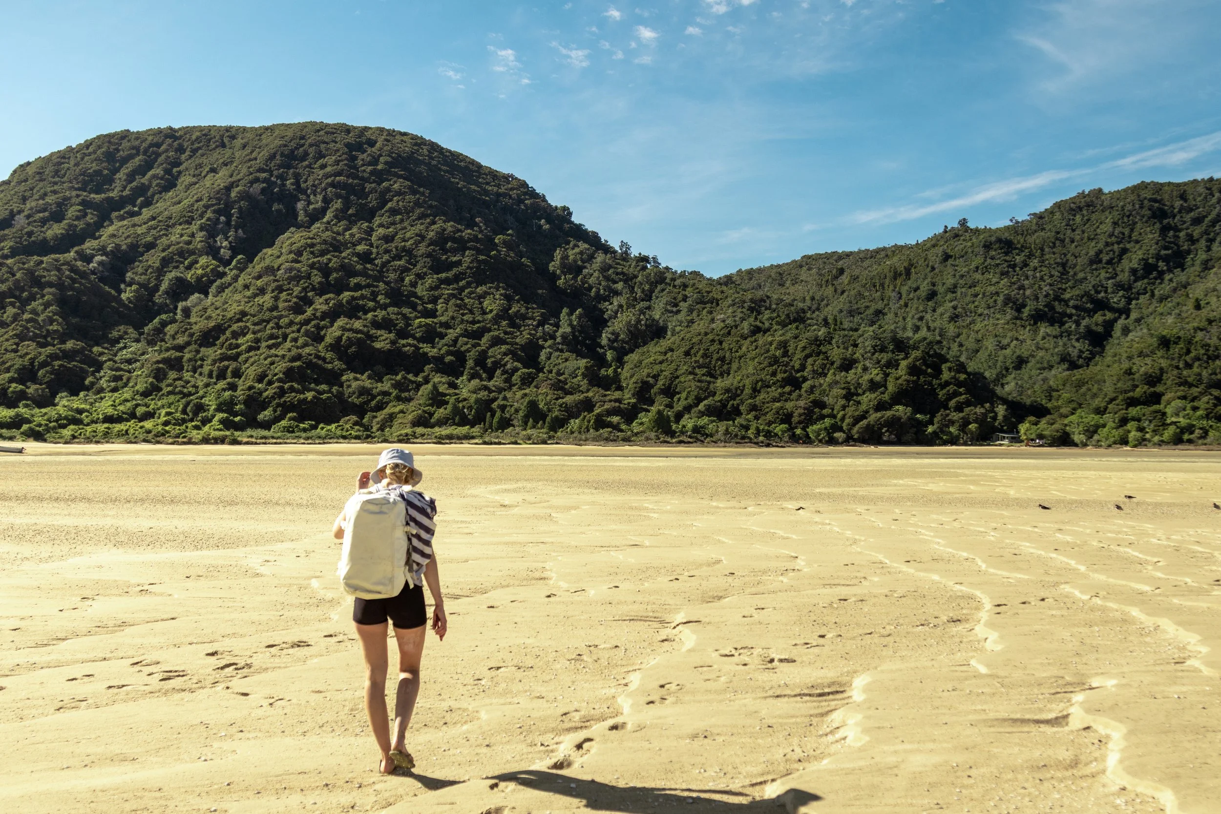 A woman with a backpack and wearing a hat walks on a sandy beach, with green hills covered in dense trees in the background and a blue sky overhead.