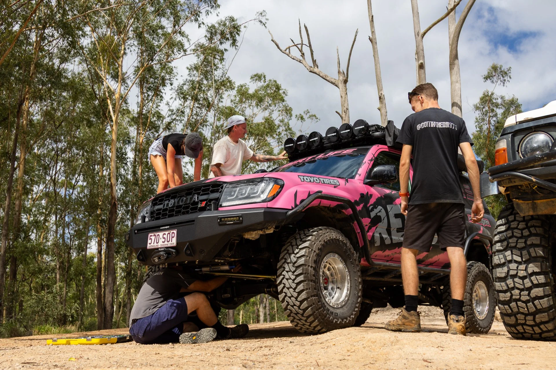 Several people work on a customized pink off-road vehicle in a wooded outdoor area. One person is underneath the vehicle, and others are around it, working on or inspecting various parts.