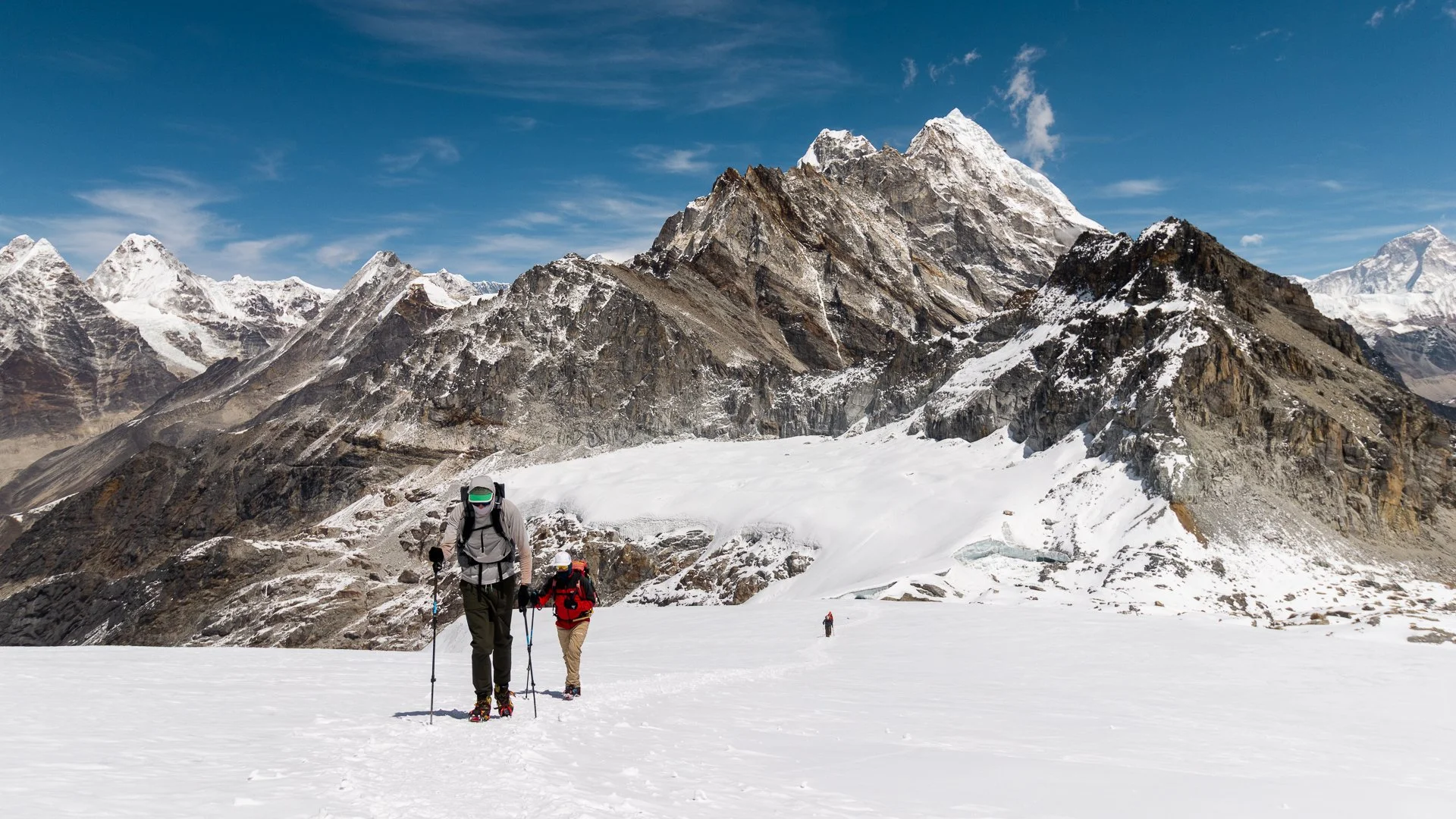 Mountains with snow and rocky peaks, a glacier, blue sky with clouds, and three hikers in winter gear trekking over snow in the foreground.
