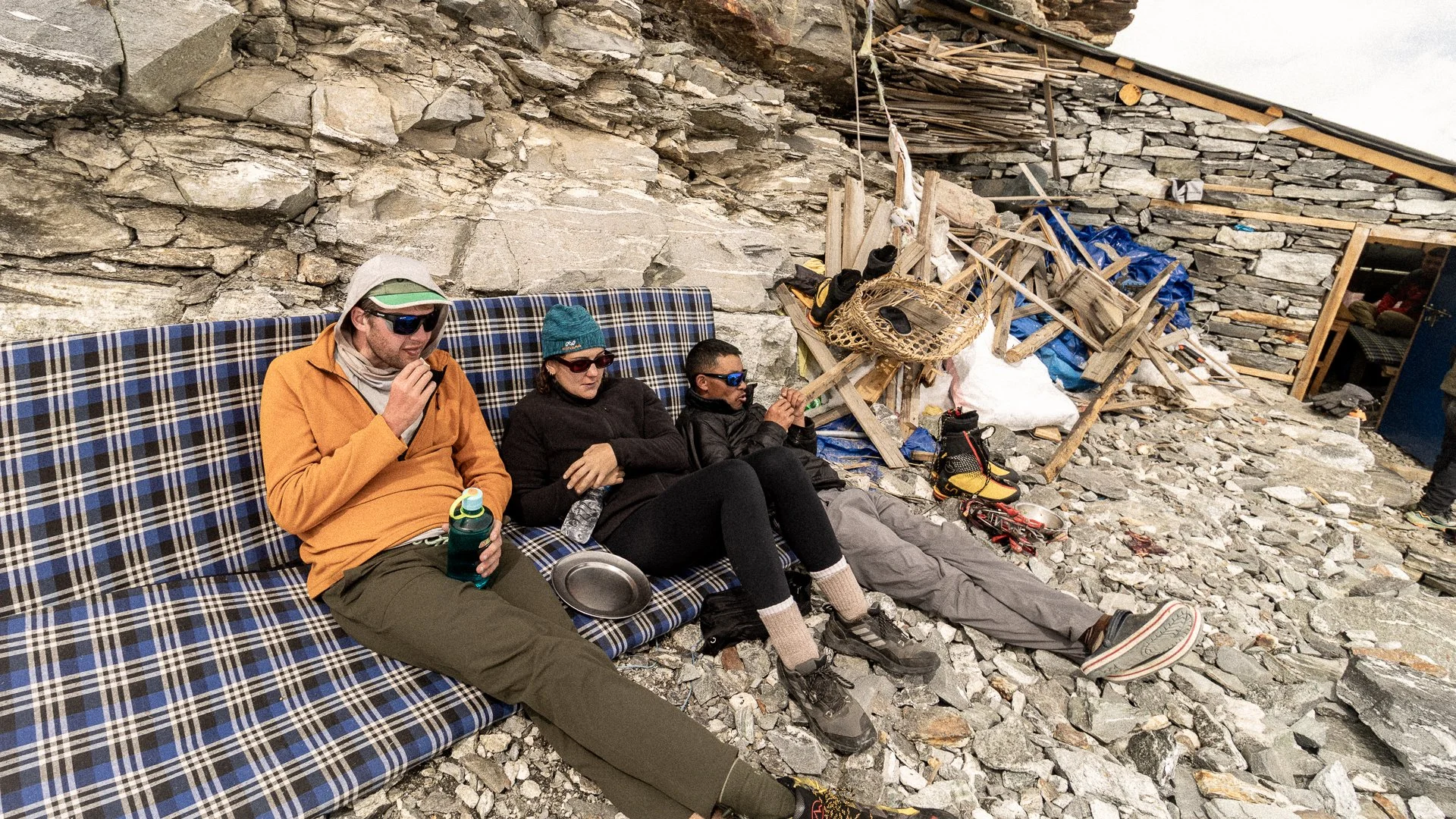Three climbers resting on a plaid-patterned bench at a rocky mountain base, with gear and makeshift shelter materials around them.