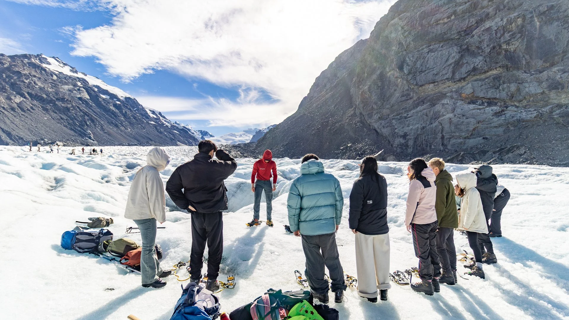 Group of people standing and sitting on snow-covered ground in a glacier landscape with mountains in the background, some wearing winter clothing and snowshoes.