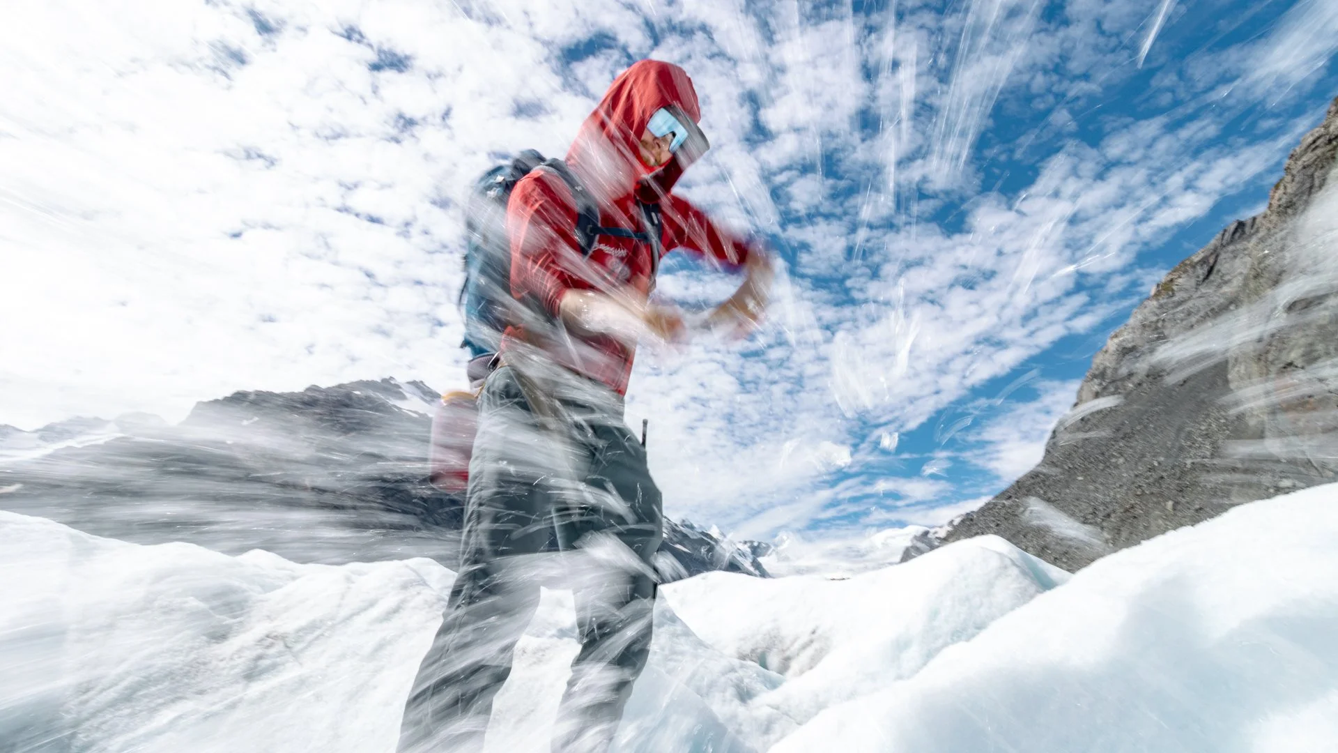 A person wearing a red jacket, snow goggles, and a backpack is kneeling in snow, surrounded by a snowy mountain landscape under a partly cloudy blue sky.