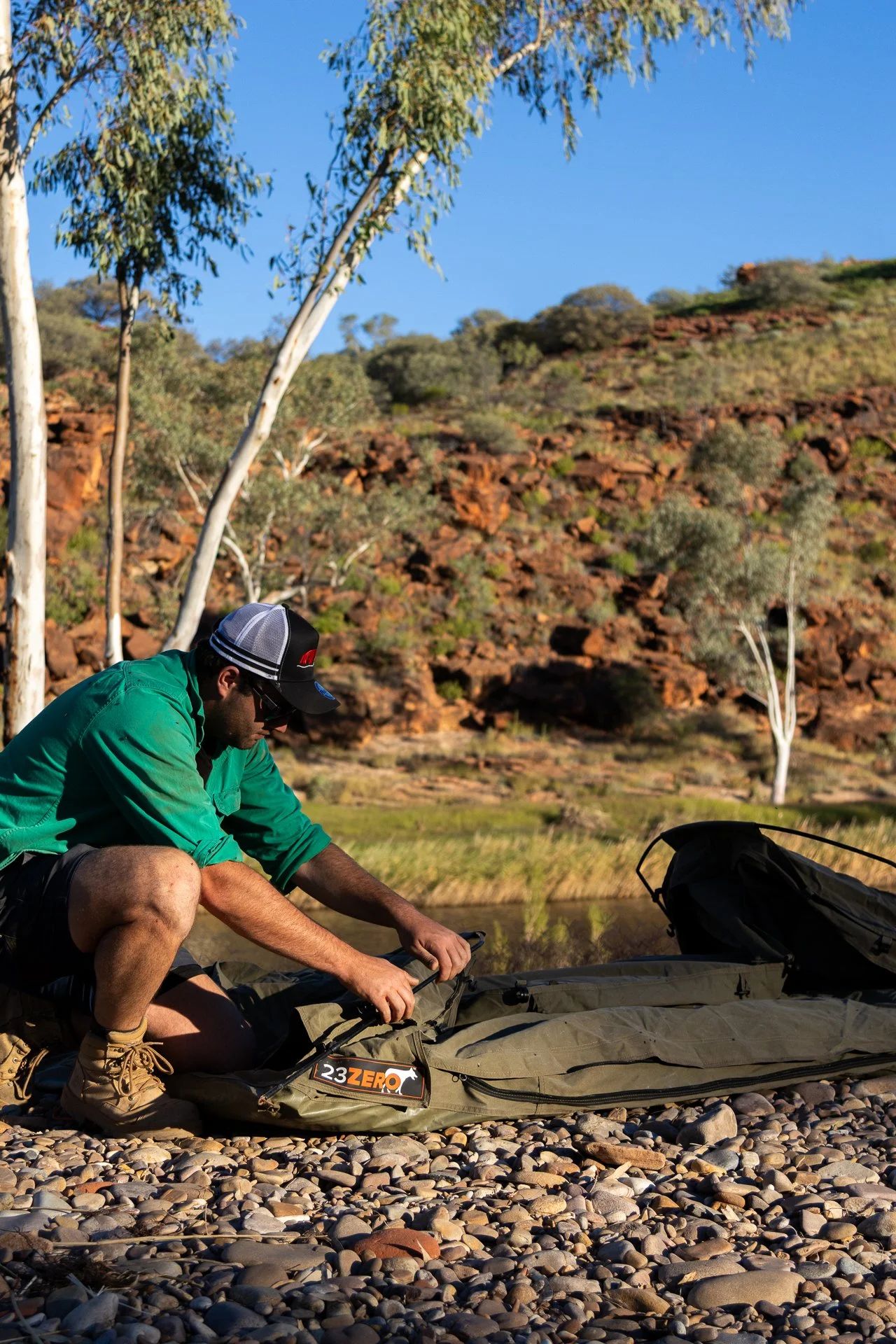 A man wearing a green jacket, baseball cap, and sunglasses kneels on a rocky riverbank, preparing a fishing rod and tackle box with a scenic landscape of trees, rocky hills, and a clear blue sky in the background.