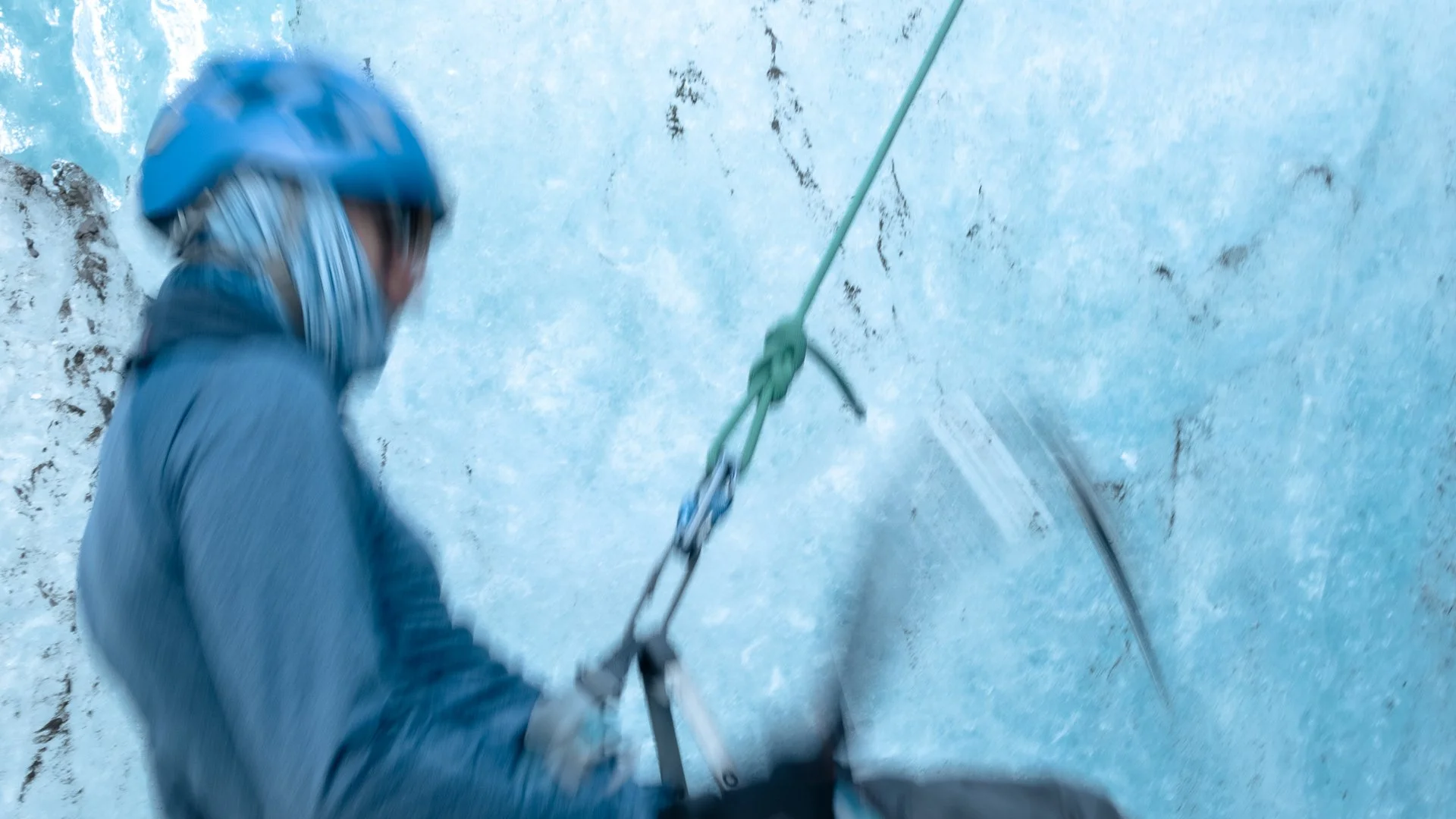 A person wearing a helmet and climbing gear is ascending an indoor ice wall with visible cracks and ice formations.