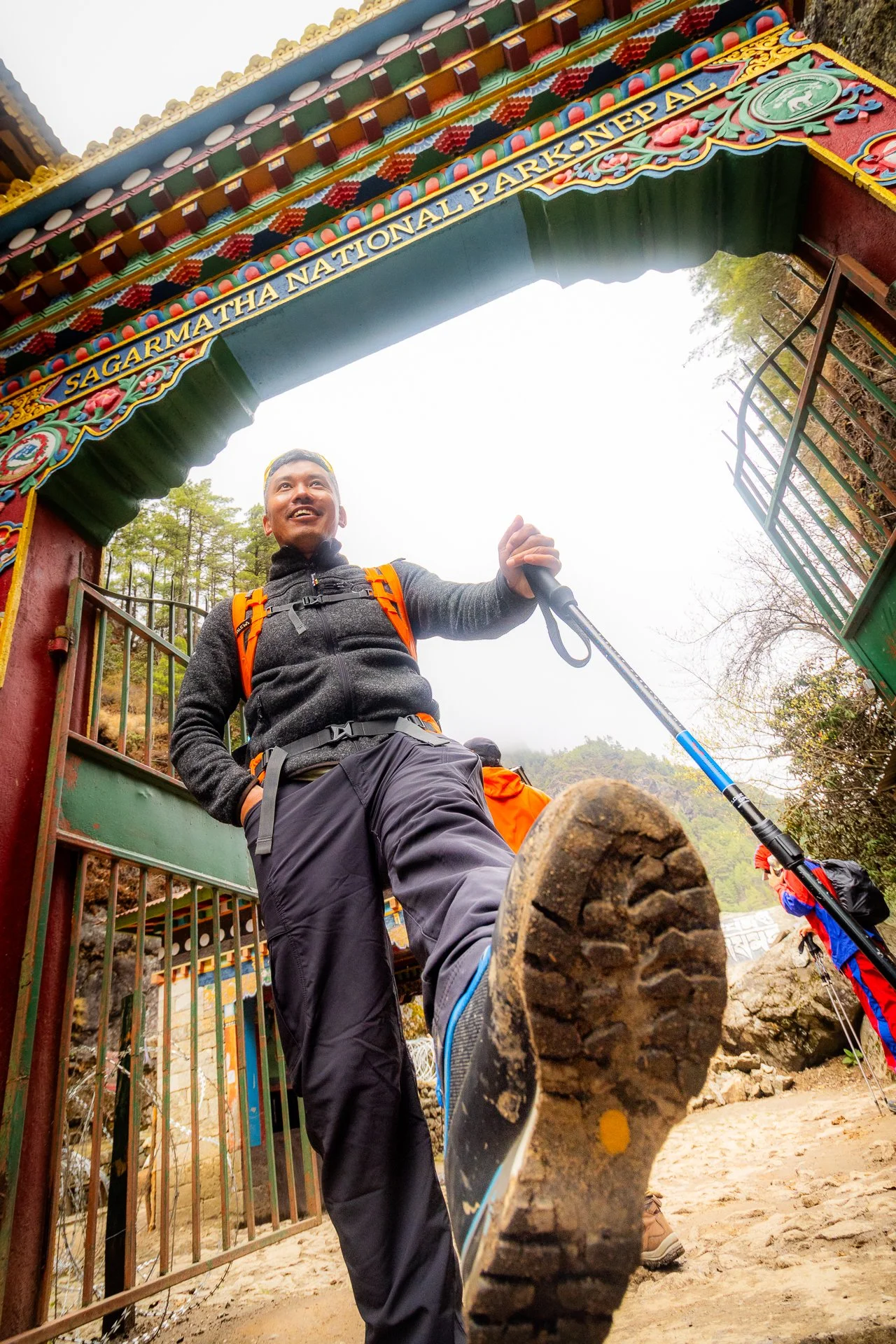 A man standing in front of an arched entrance to Sagrada Thá National Park in Nepal, smiling and holding a hiking pole, with mud on his hiking shoe, and others in the background