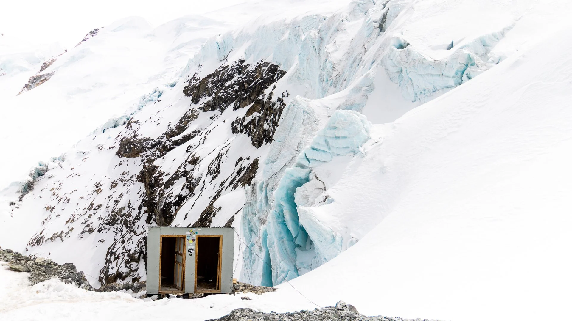 A small structure with open doors is situated in a snowy, mountainous landscape with large ice formations and snow-covered slopes.