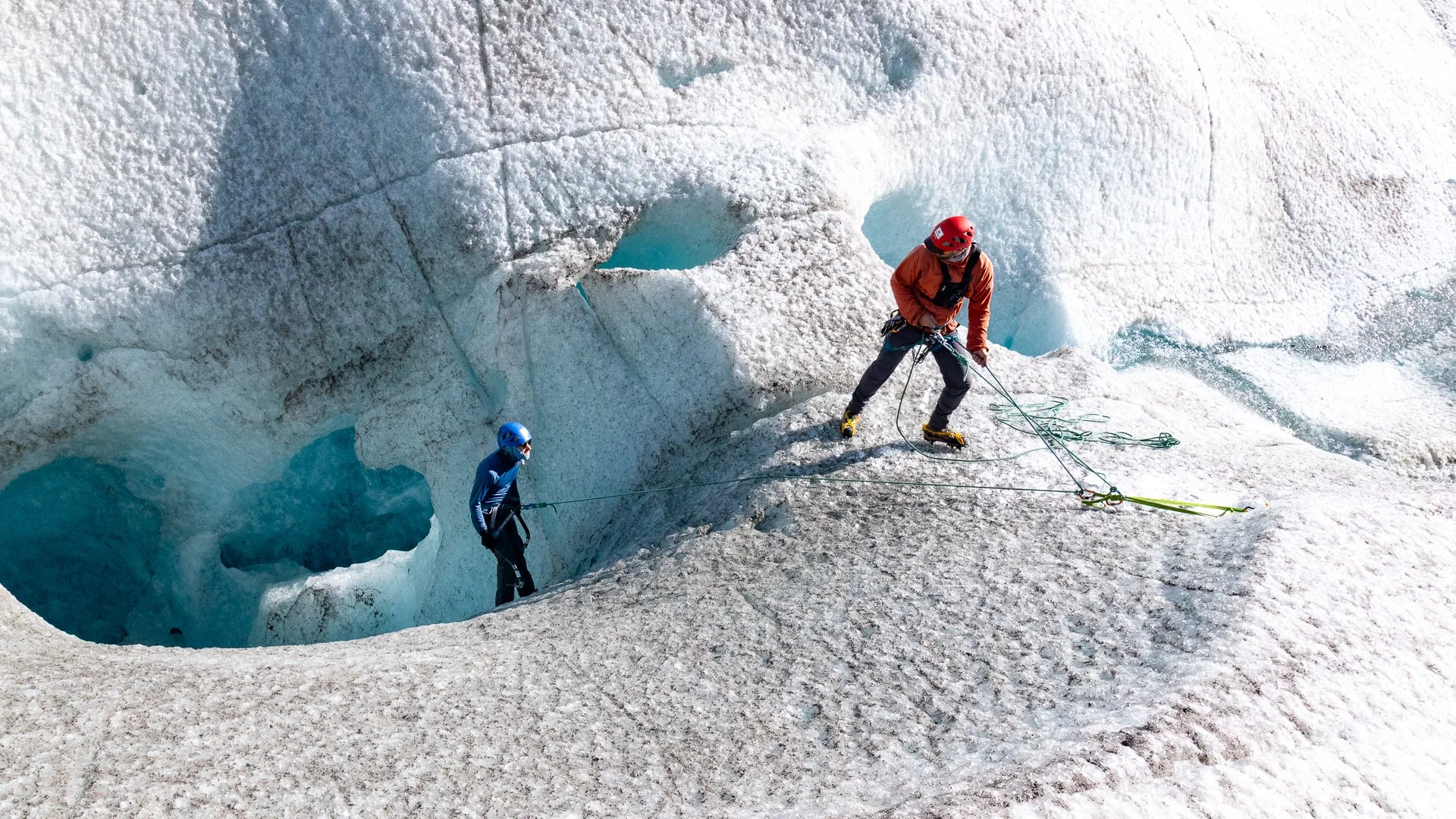 Two climbers on an icy glacier, one wearing a red jacket and yellow crampons, the other in a blue jacket, both secured with ropes and wearing helmets.