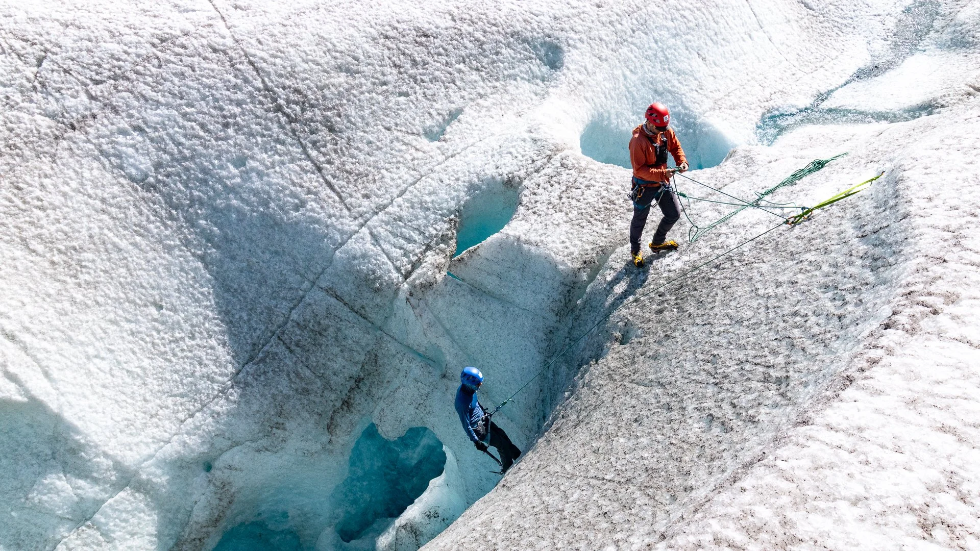 Two climbers wearing helmets, harnesses, and cold weather gear ascending and descending on a crevasse in a glacier with snow and ice.