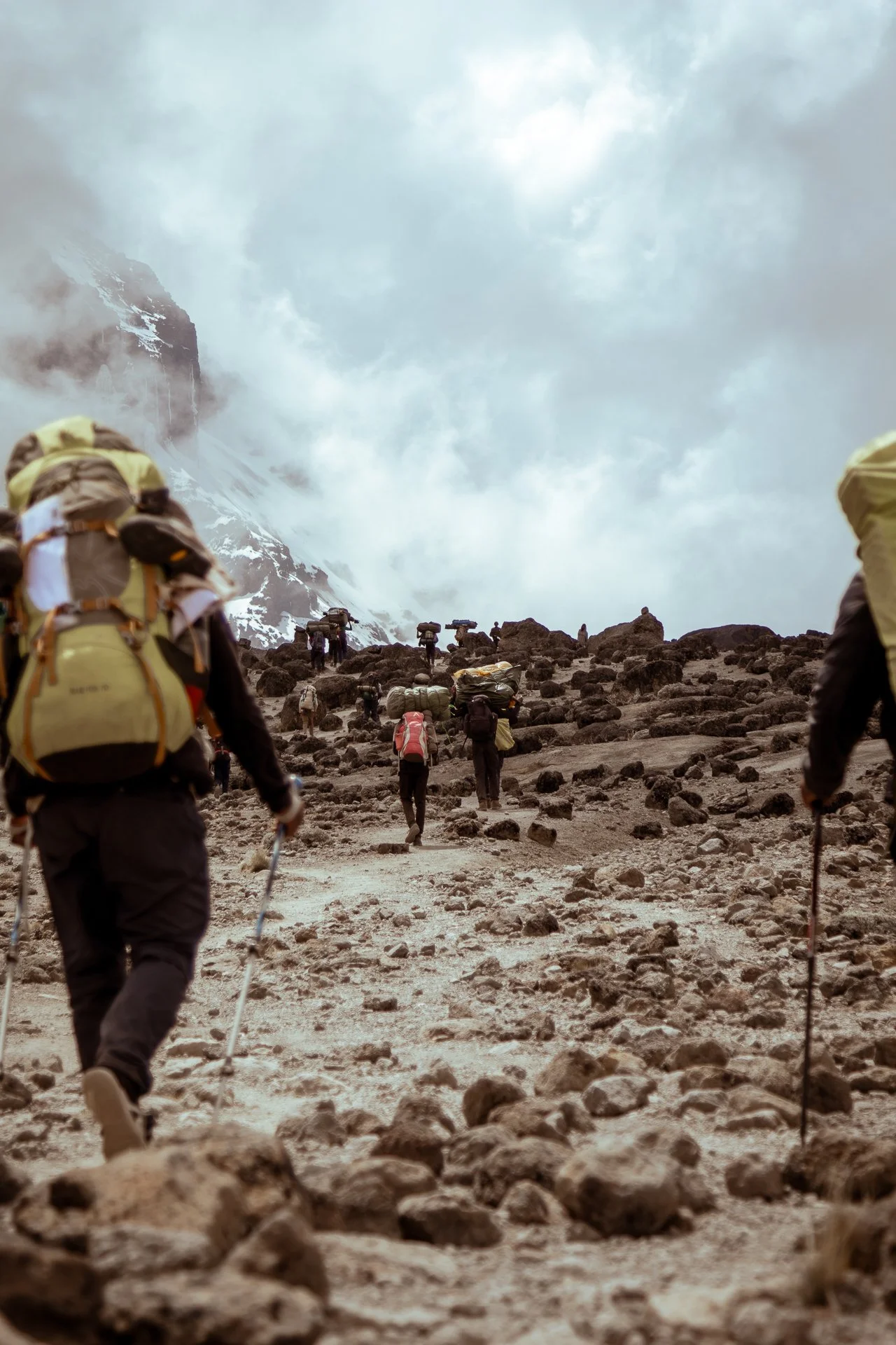 Group of hikers walking on rocky trail in mountainous, cloudy terrain.