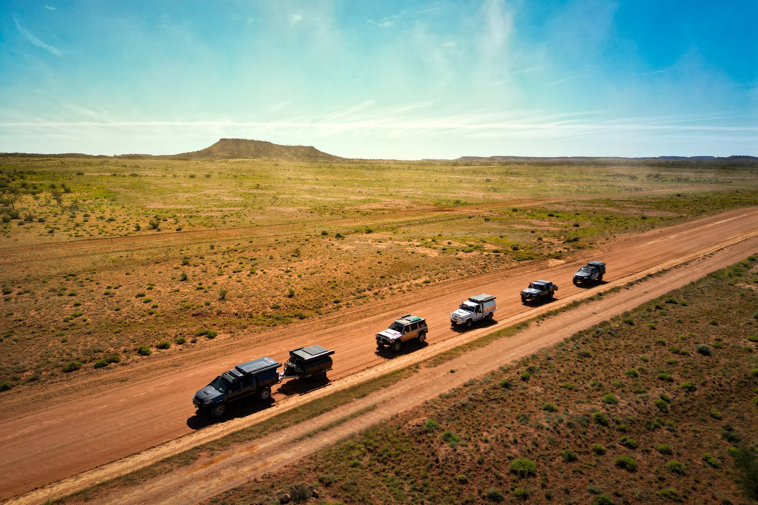 An aerial view of a desert landscape with a dirt road along which several off-road vehicles are driving. The scene features sparse vegetation, a flat-topped mountain in the background, and a clear blue sky.