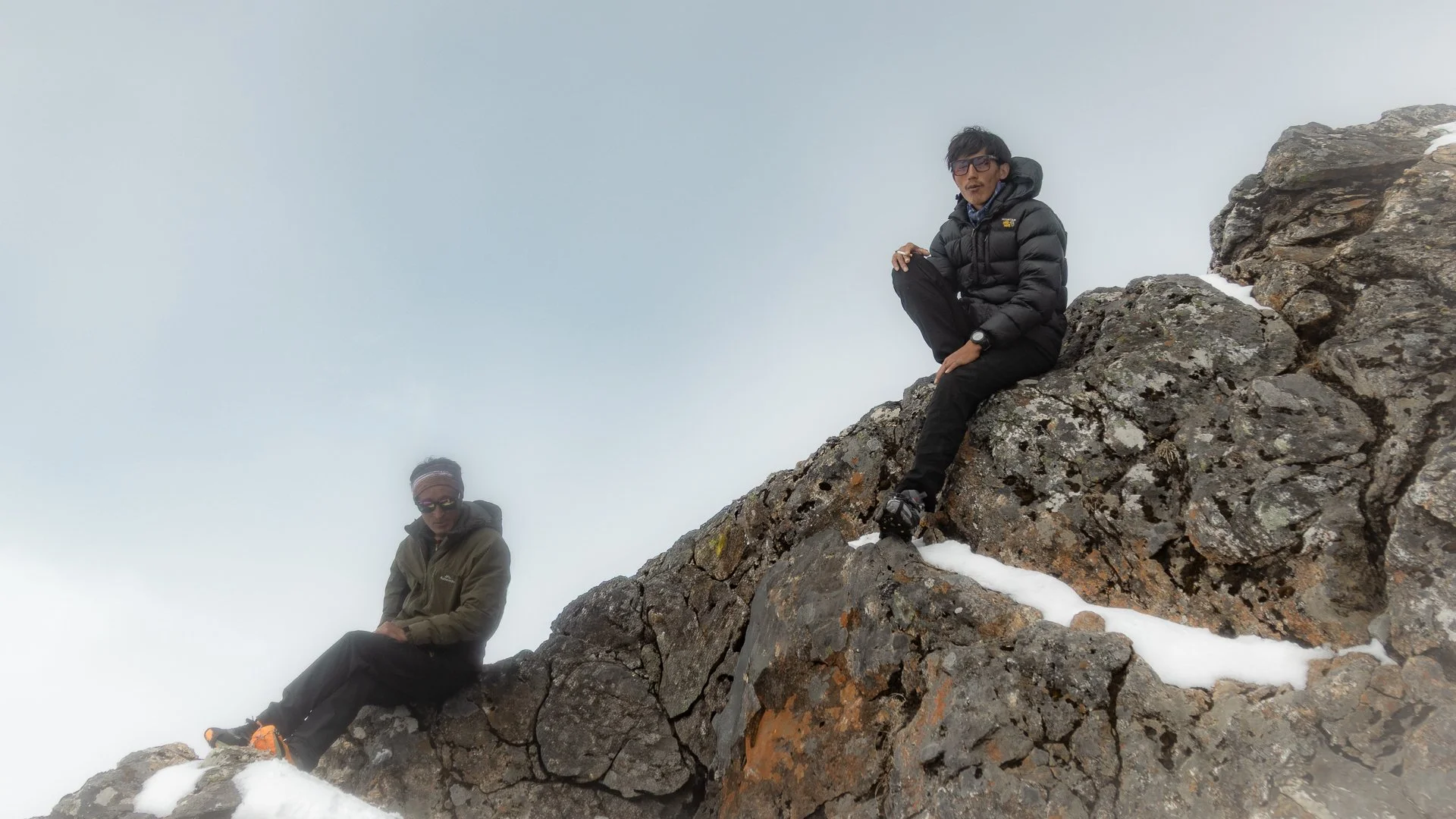 Two people sitting on a rugged, snow-dusted mountain slope in a foggy, high-altitude environment.