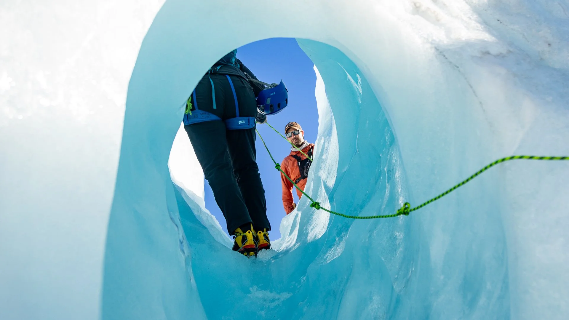 Two mountaineers in winter gear inside an ice cave, connected by a green rope, with one ascending and the other watching, on a bright sunny day.
