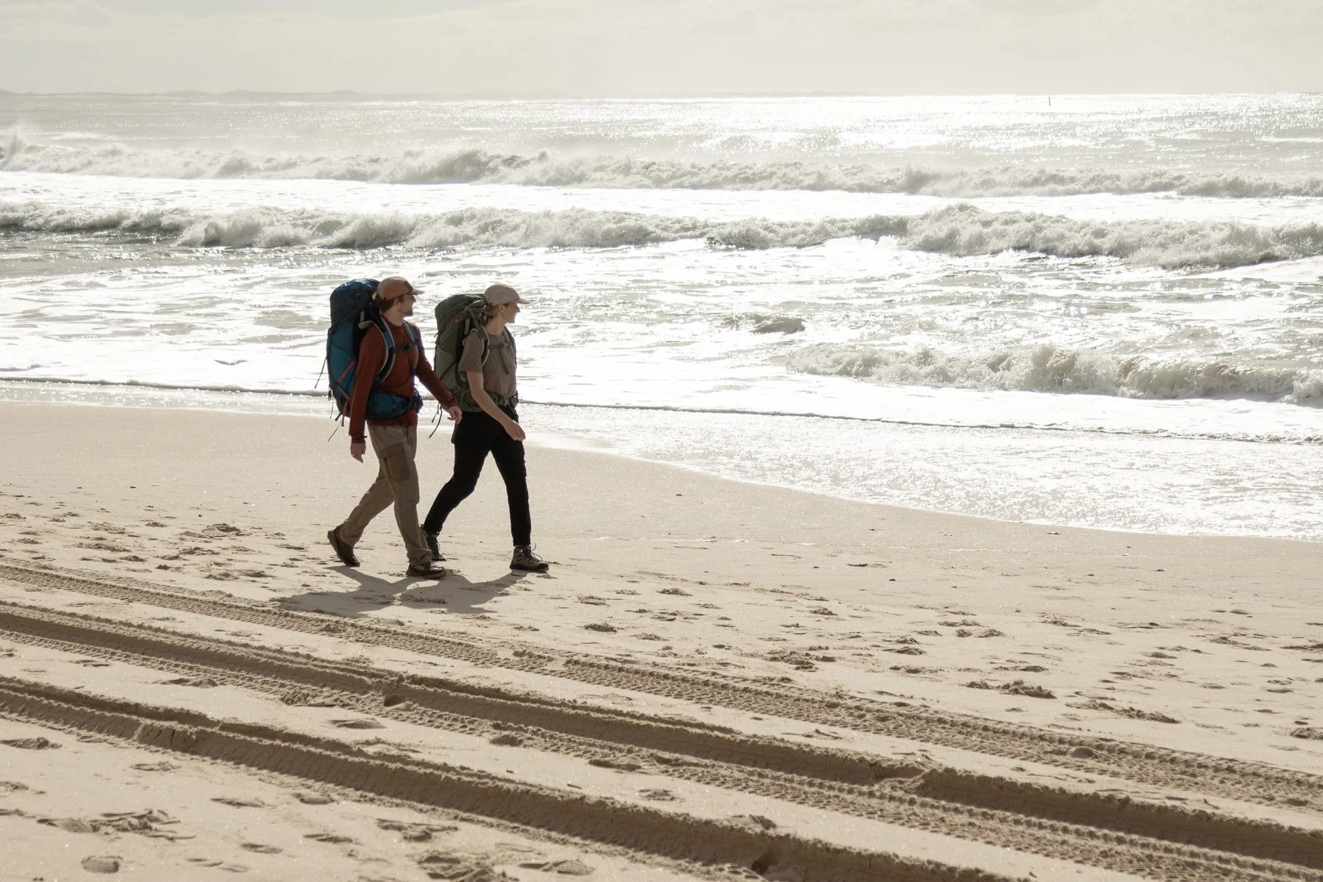 Two people with backpacks walking along a sandy beach near the ocean, tire tracks visible in the sand, overcast sky with sunlight reflecting off the water.