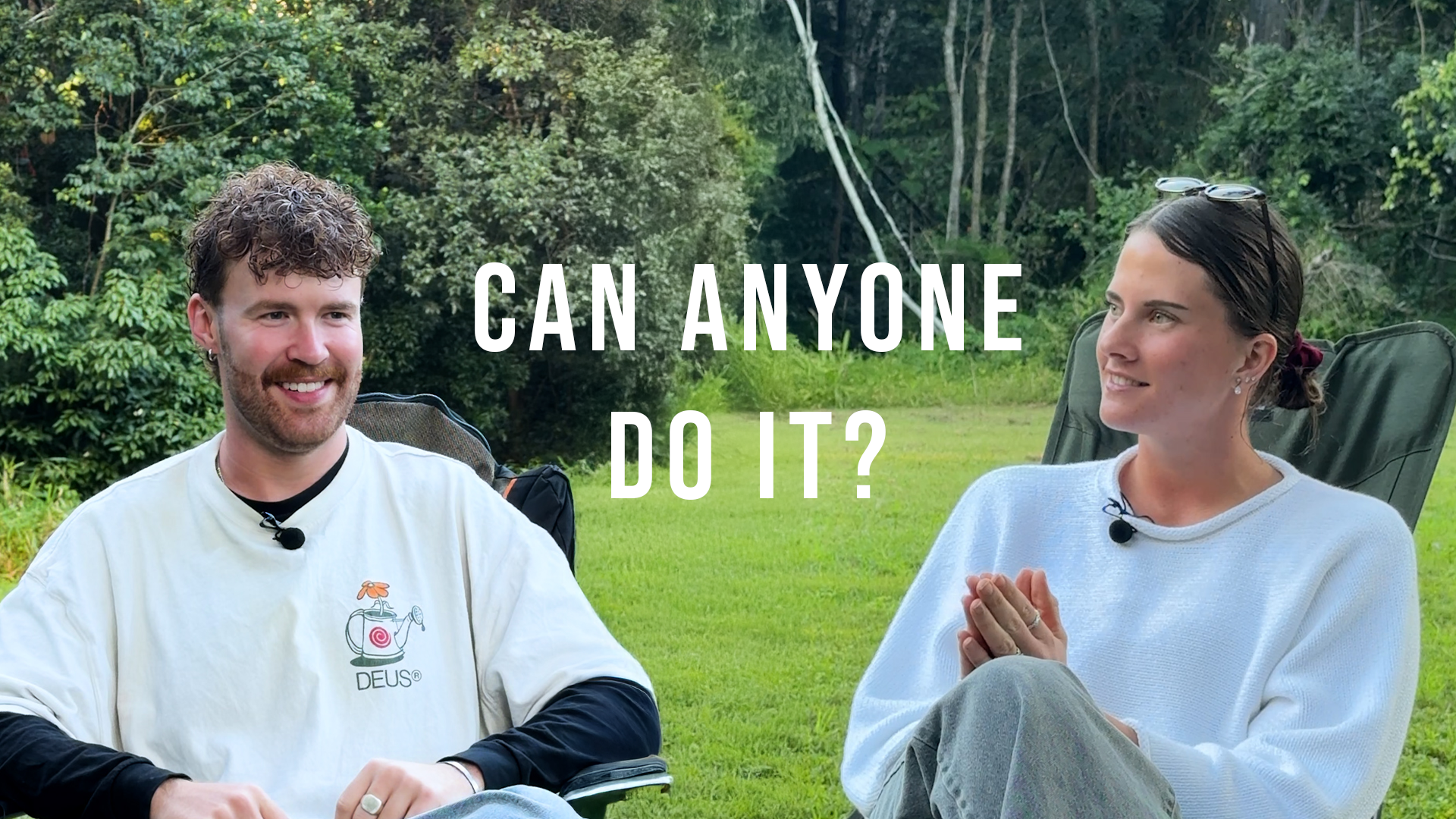 Two people sitting outdoors on grass, engaged in a conversation with text overlay asking 'Can anyone do it?' The man on the left has curly hair, a beard, a white shirt with a logo, and is smiling. The woman on the right has dark hair tied back, sunglasses on her head, and is gesturing with her hands. They are surrounded by green trees and foliage.