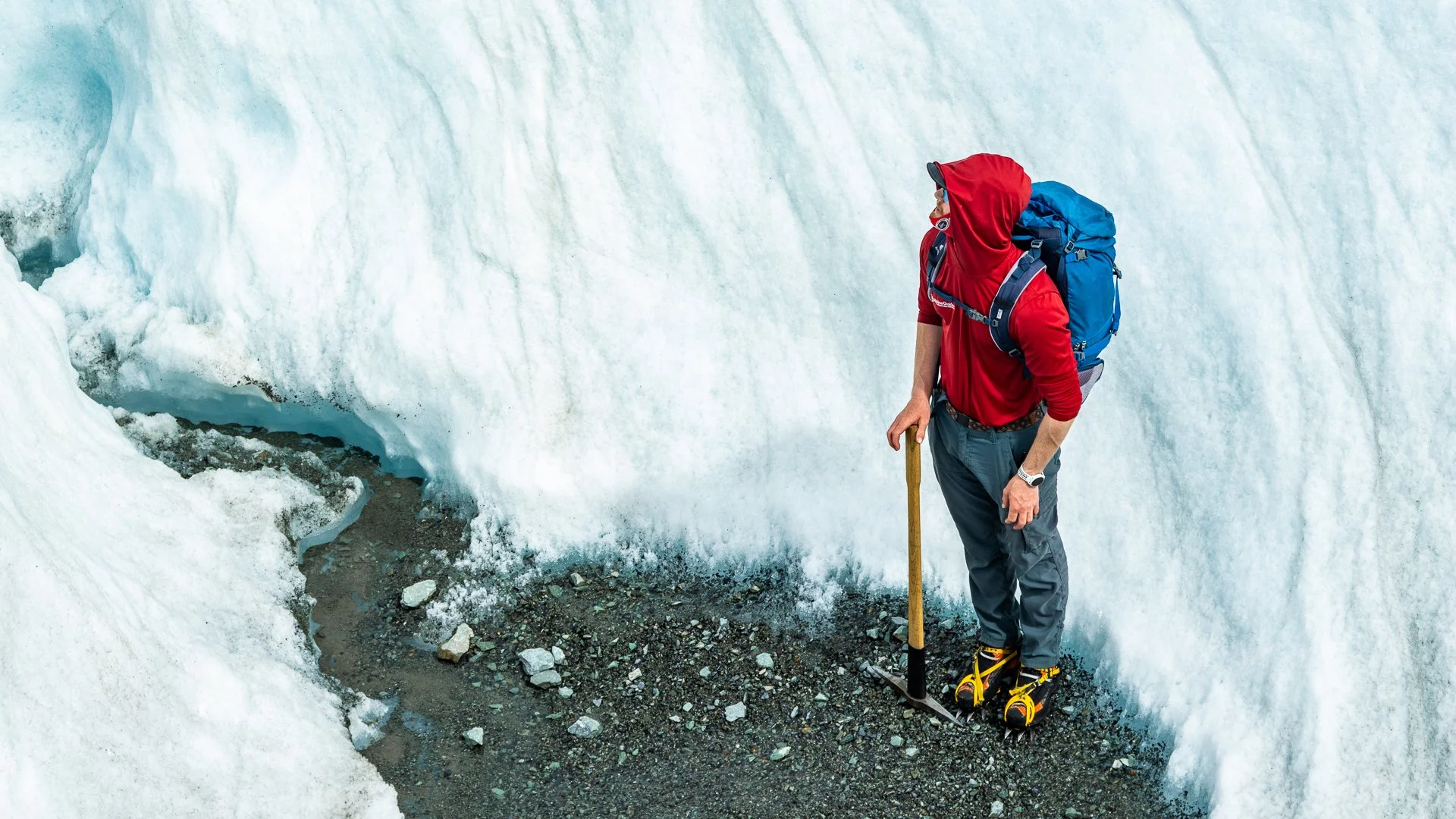 A man in a red hoodie, gray pants, and hiking boots with crampons, standing on rocky terrain next to a large snow glacier, holding a walking stick.