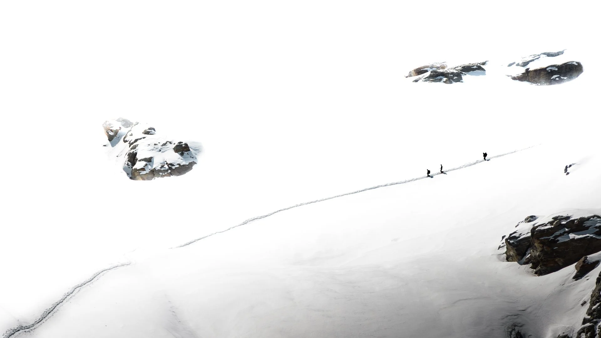 Three hikers walking in single file across a snowy mountain landscape with large rocks and snow-covered terrain.
