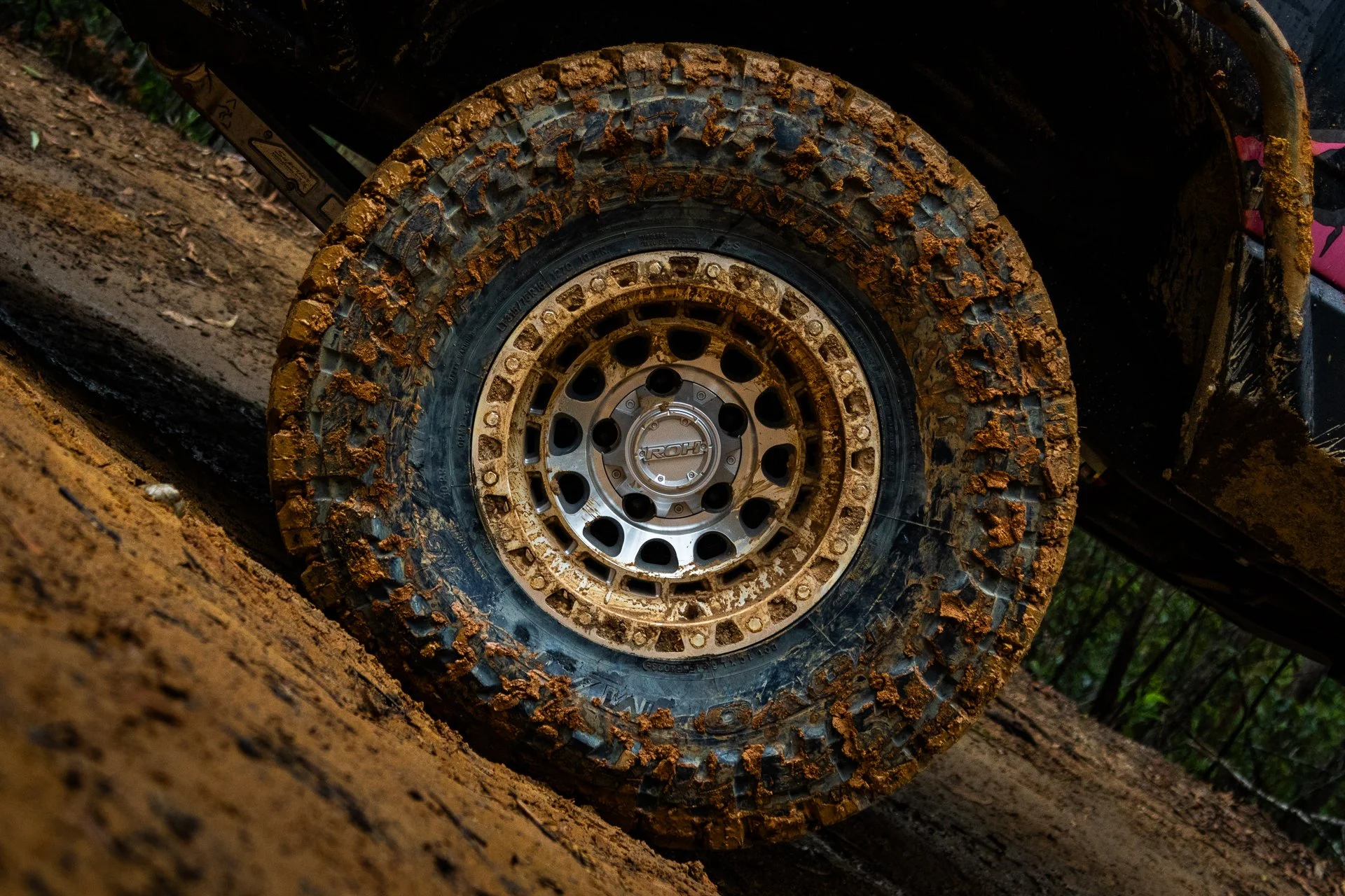 Close-up of a rugged off-road vehicle tire coated with mud, attached to a black vehicle on a dirt trail.