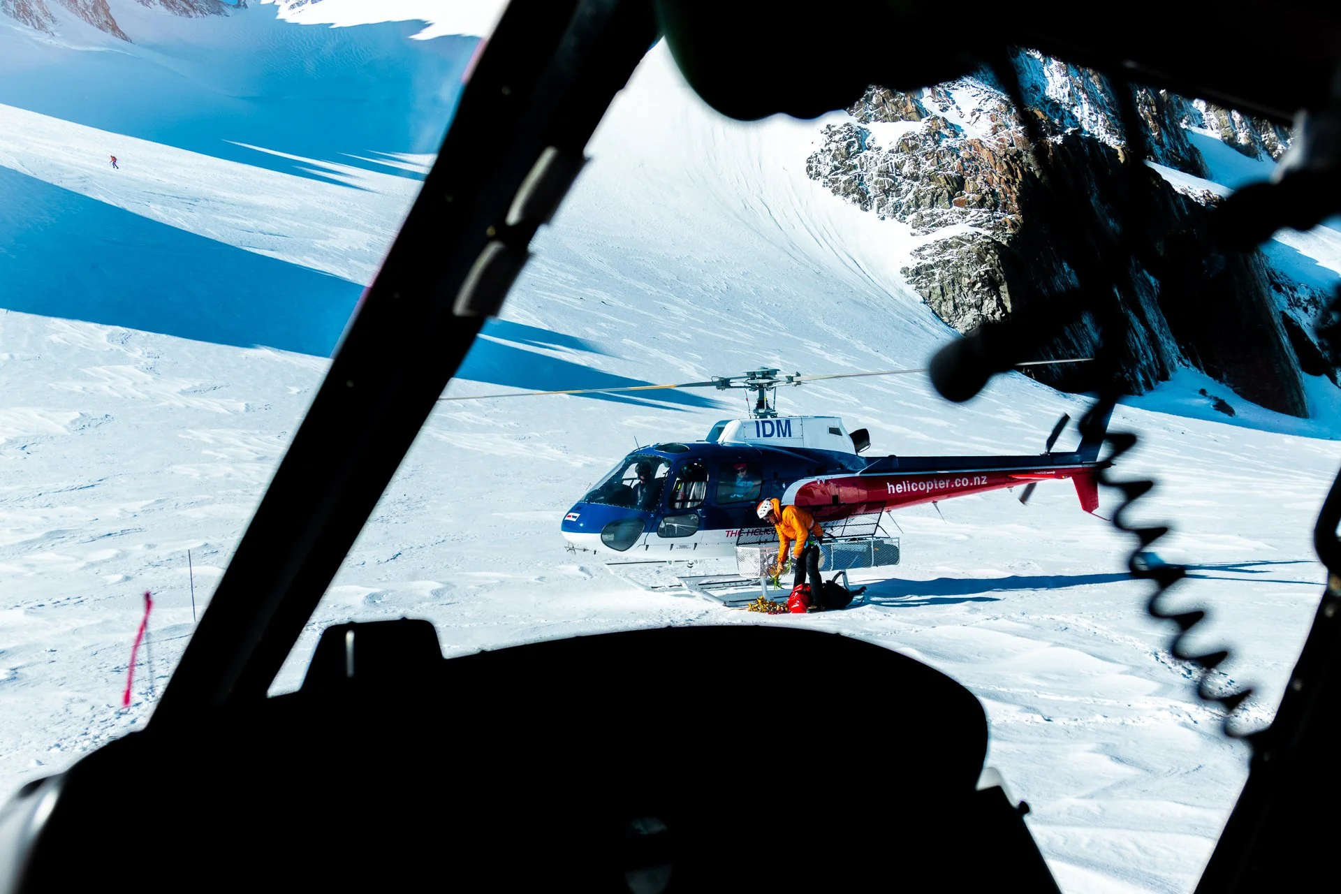 View from inside a helicopter cockpit looking out at a snowy mountain landscape with a helicopter on the ground and a person in orange gear near the helicopter.