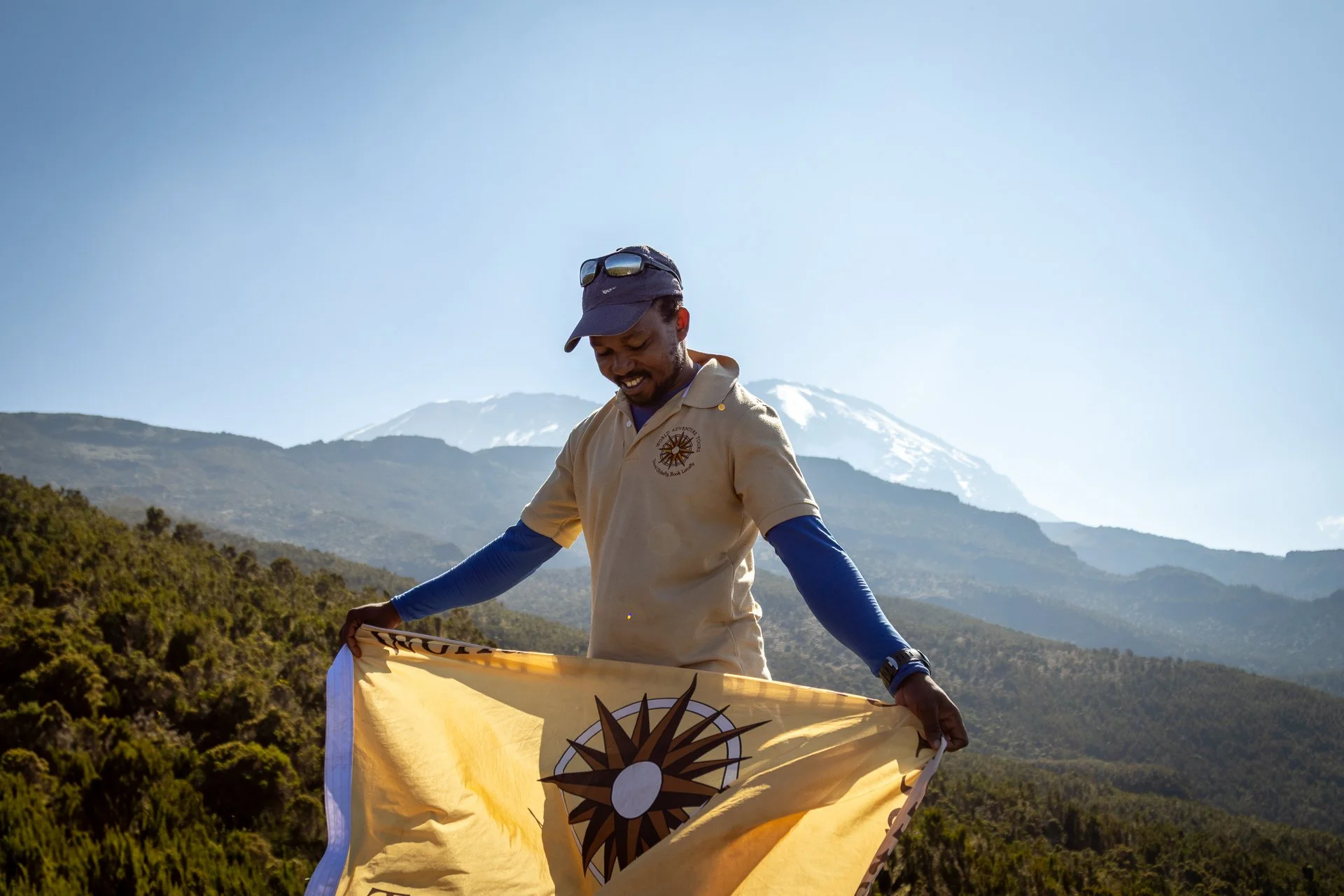 A man in beige shirt holding a yellow flag with a sunburst emblem, standing outdoors with mountains and a snow-capped volcano in the background.
