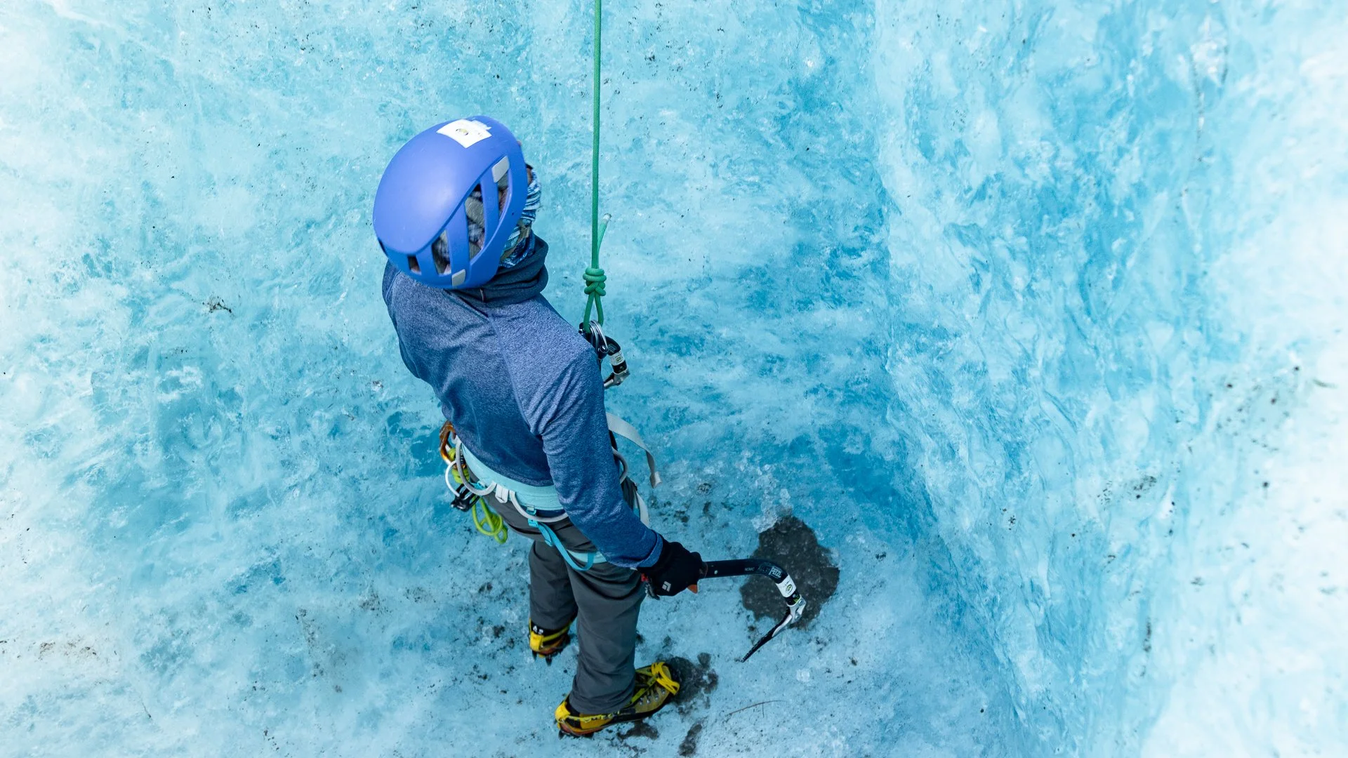 A person wearing a blue helmet and winter gear ice climbing on a steep icy wall, holding ice axes.