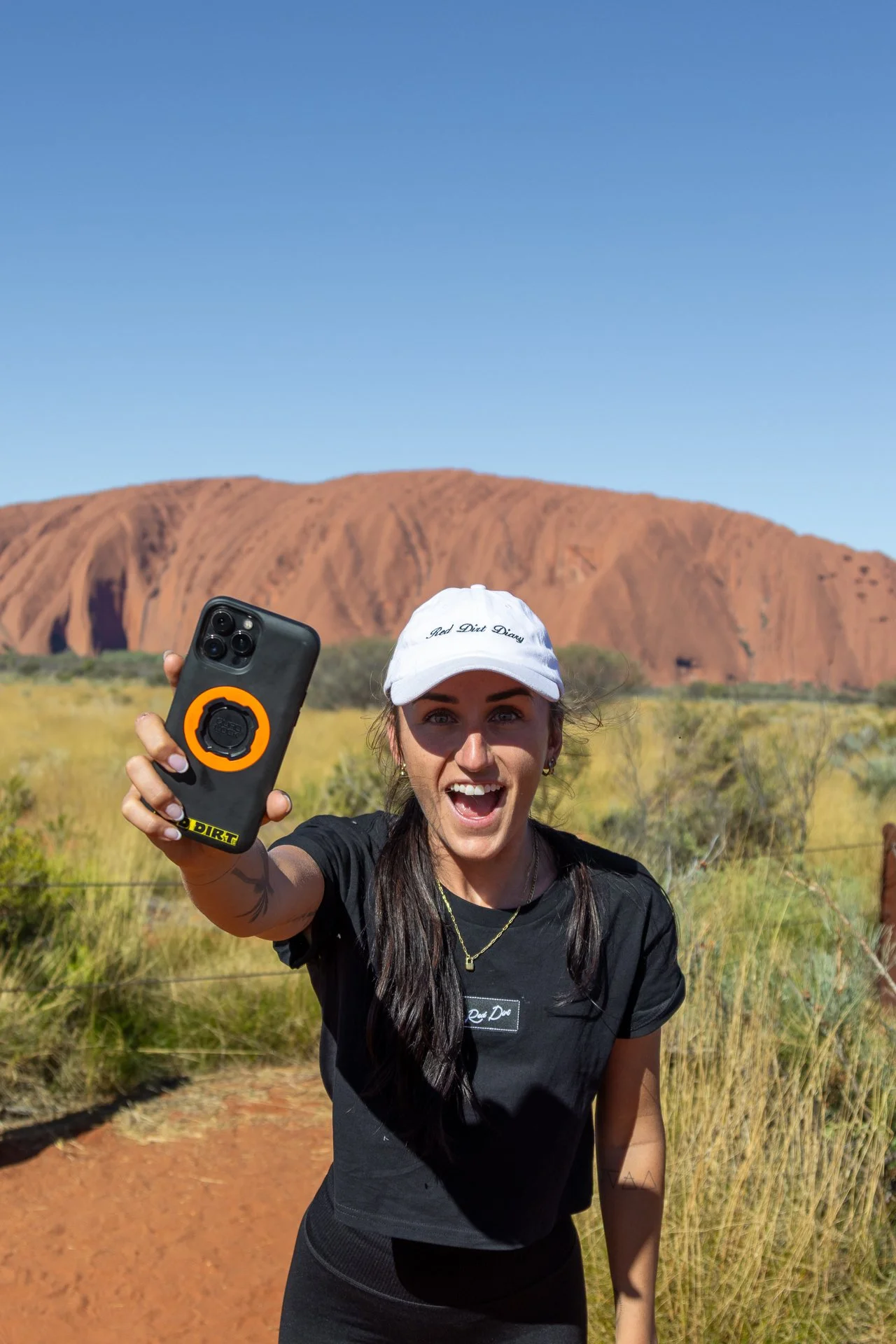 A woman smiling and taking a selfie with her smartphone in a sunny outdoor area, with a large red rock formation and blue sky in the background.
