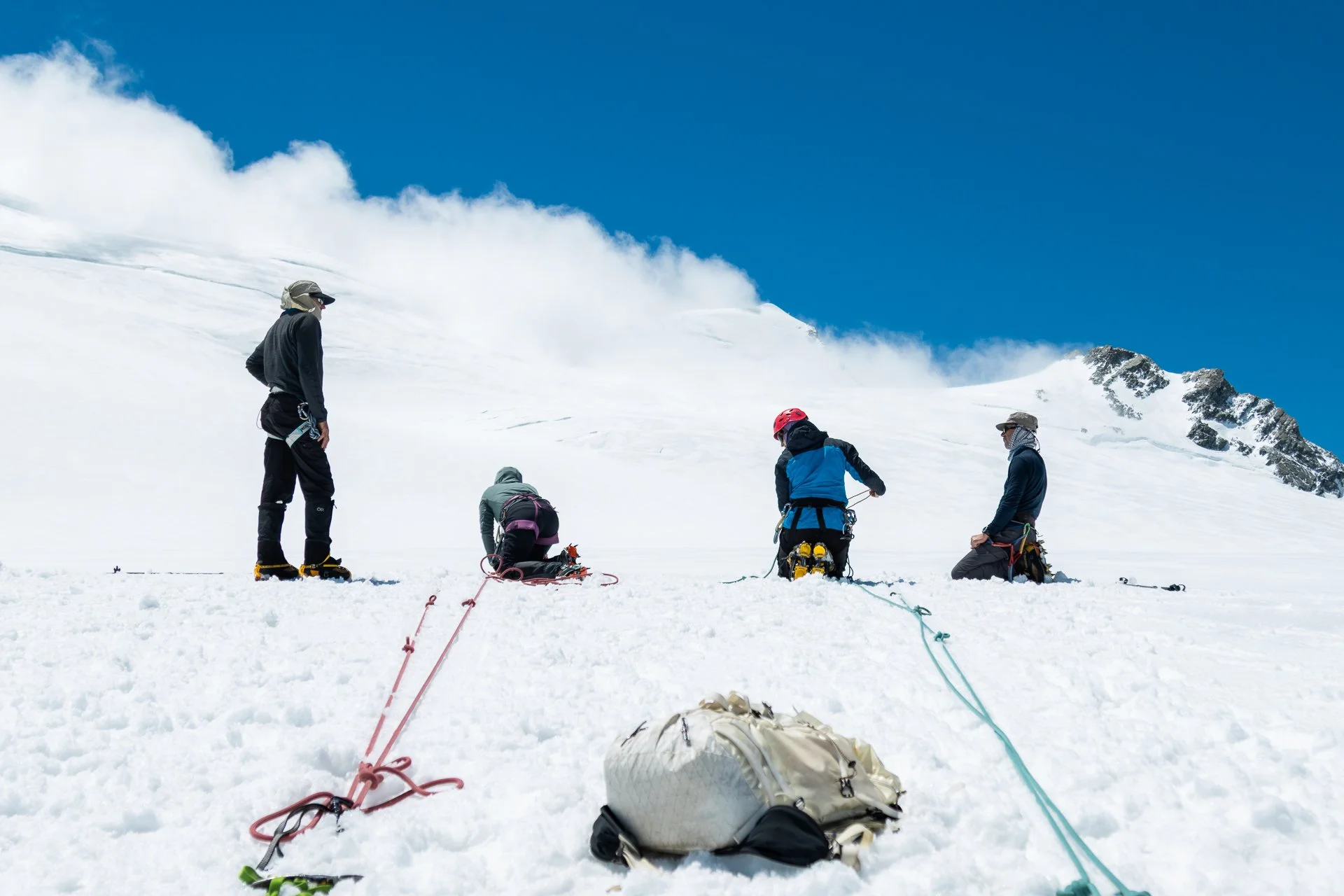 Group of climbers on snow-covered mountain preparing for ascent, with ropes, backpacks, and gear, under a blue sky and surrounded by snow and rocky peaks.