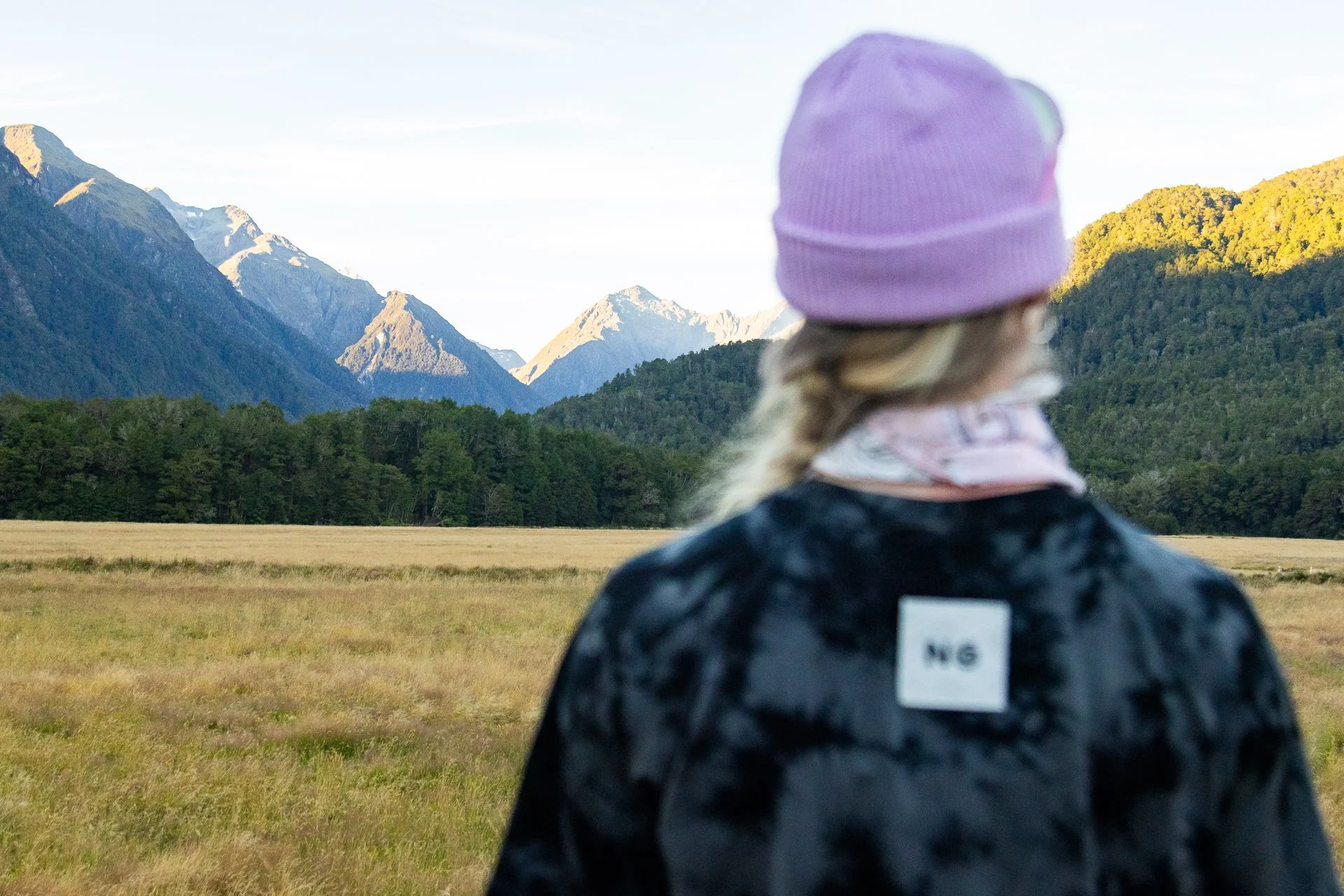 Person wearing a purple knit hat and a dark jacket with a white label on the back, looking at a mountain landscape with green forested hills and snow-capped peaks in the distance.